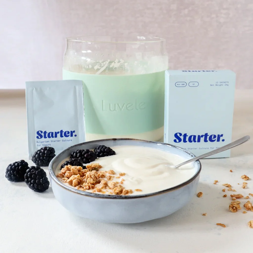 A bowl of yogurt with granola and blackberries, a spoon inside, with a glass of milk, and boxes of starter culture packets in the background.