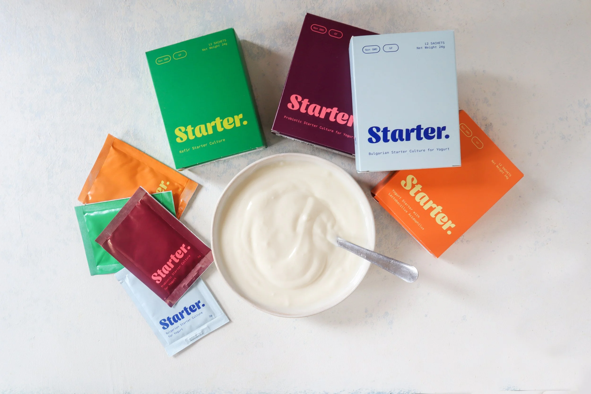 Assorted colorful starter culture boxes and a bowl of yogurt on a white surface.