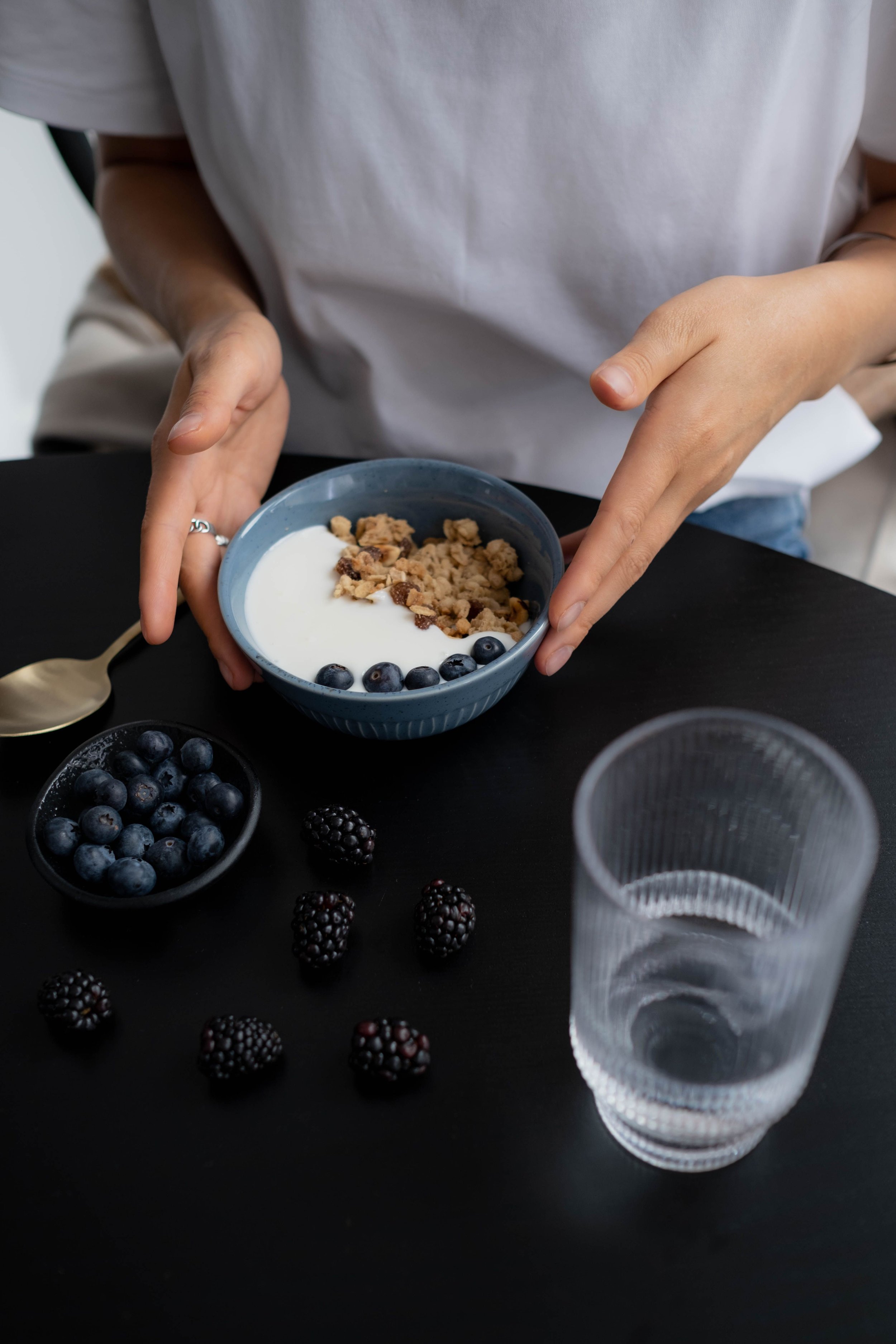 A person holding a bowl of yogurt topped with granola and blueberries, with additional blueberries and blackberries on a black table, and a clear empty plastic cup.