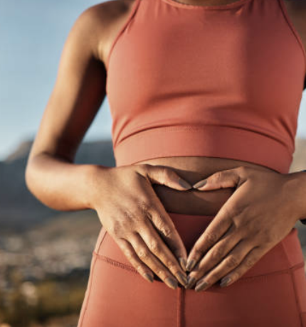 A woman wearing a sports bra and leggings making a heart shape with her hands over her stomach.