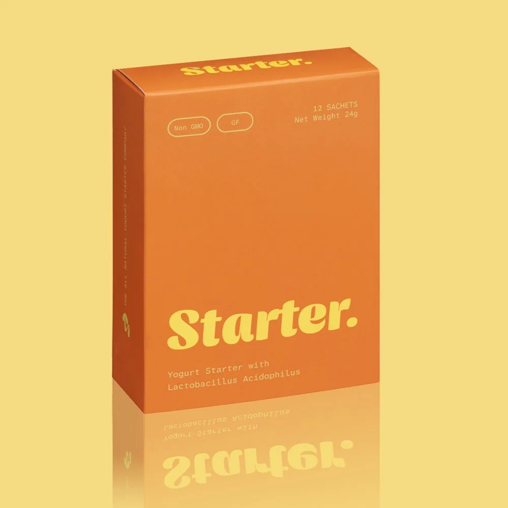 Orange box of yogurt starter with lactose acidophilus, labeled 'Starter' and indicating 12 sachets, non-GMO, gluten-free, with a reflection on a yellow background.