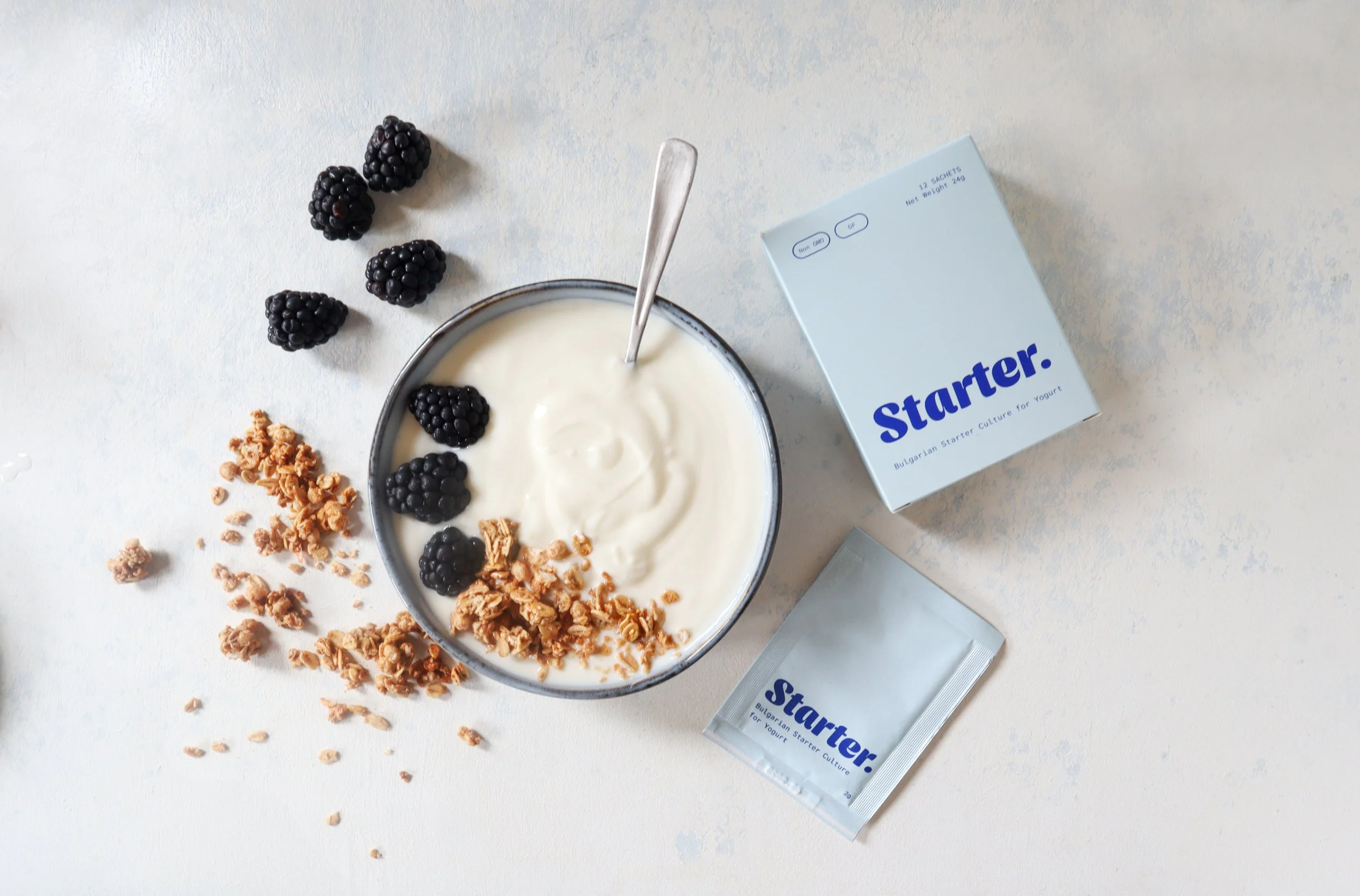 Bowl of yogurt with blackberries and granola, with a box labeled 'Starter,' on a light-colored surface.