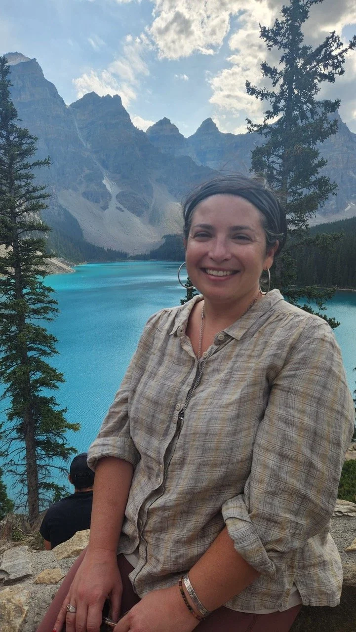 A woman smiling in front of a turquoise lake with mountains and trees in the background.