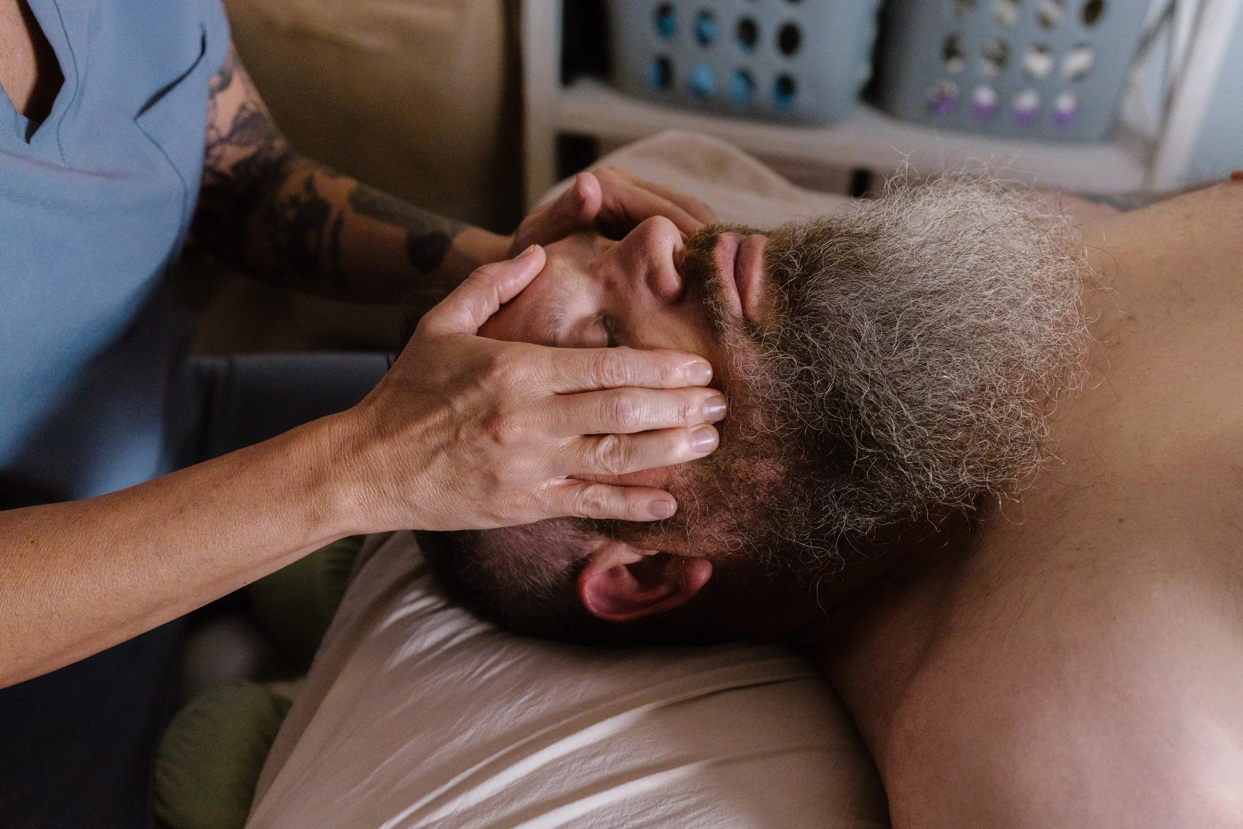 A caregiver is giving a facial massage to a man with a gray beard who is lying down with his eyes closed.