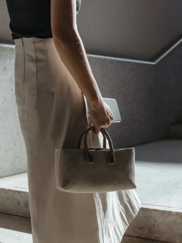 A person in beige pants and a black shirt holding a small beige handbag and a smartphone.