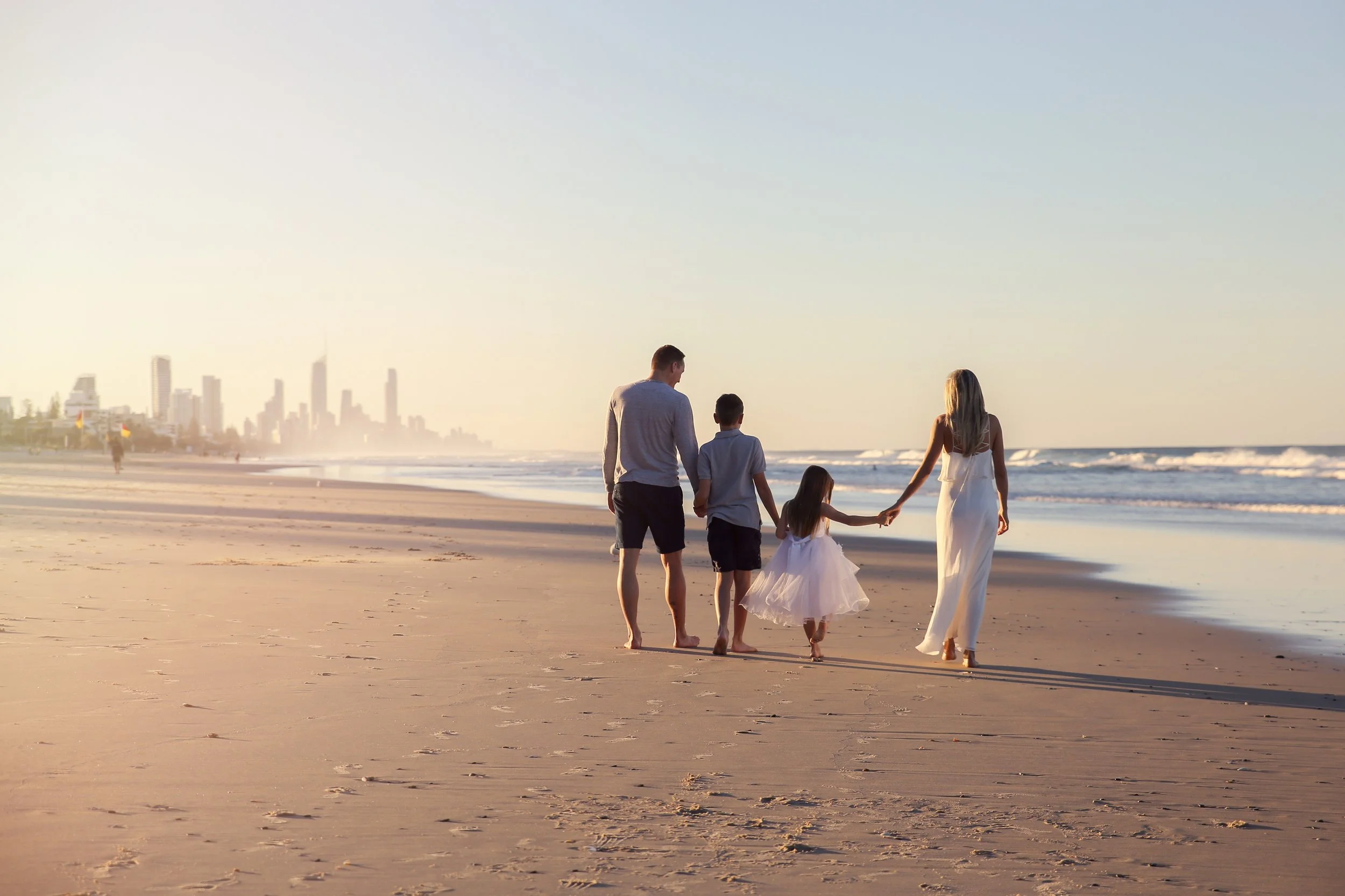 A family of five walking hand in hand on the beach during sunset with a city skyline in the background.