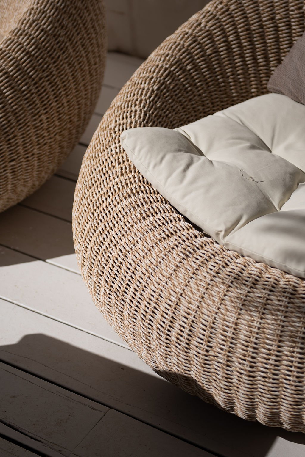 Close-up of a rattan outdoor chair with a beige cushion and a pillow on top, placed on a wooden deck with sunlight casting shadows.