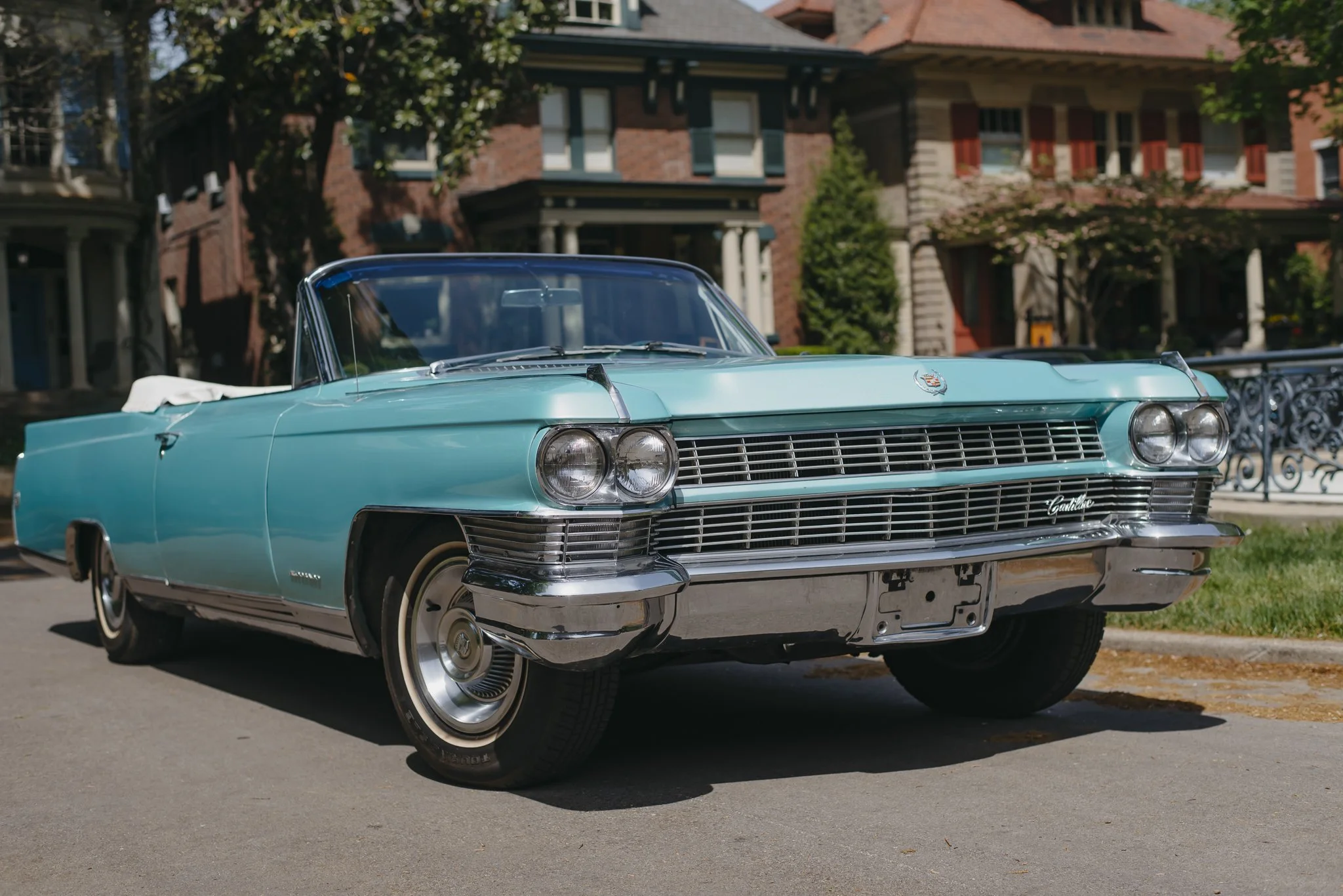 A woman with long blonde hair sitting in Charlene, a 1964 Cadillac Eldorado convertible in Turino Turquoise, wearing a large floral headpiece and looking at the camera. The background includes a lawn and a house with steps leading up to the entrance.