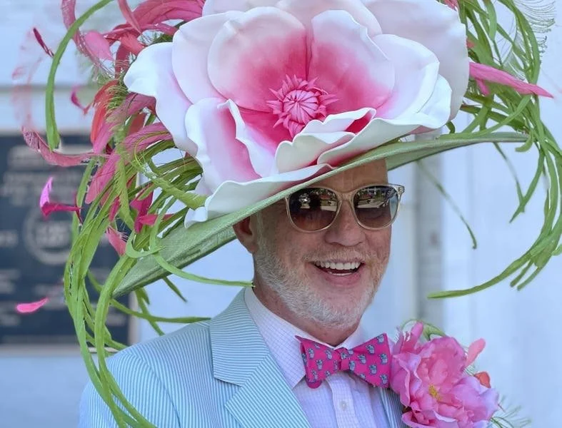 A man wearing sunglasses, a light blue striped blazer, and a pink bow tie, smiling at the Kentucky Oaks. He has a large floral hat made of pink and white flowers, with green leaves and pink accents.