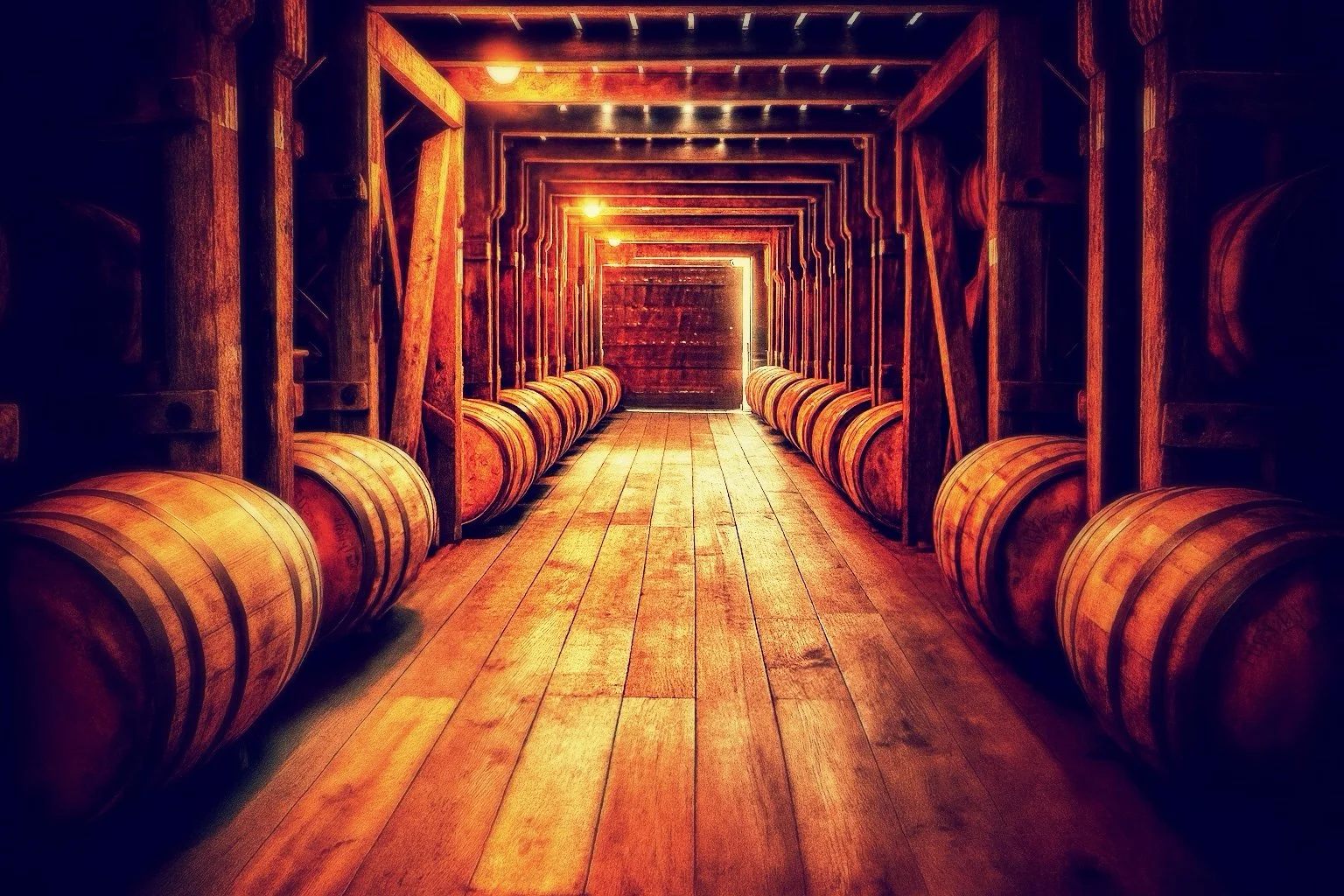 A narrow wooden walkway in a distillery rickhouse with barrels lining both sides, illuminated by warm lights, leading to a bright doorway at the end.