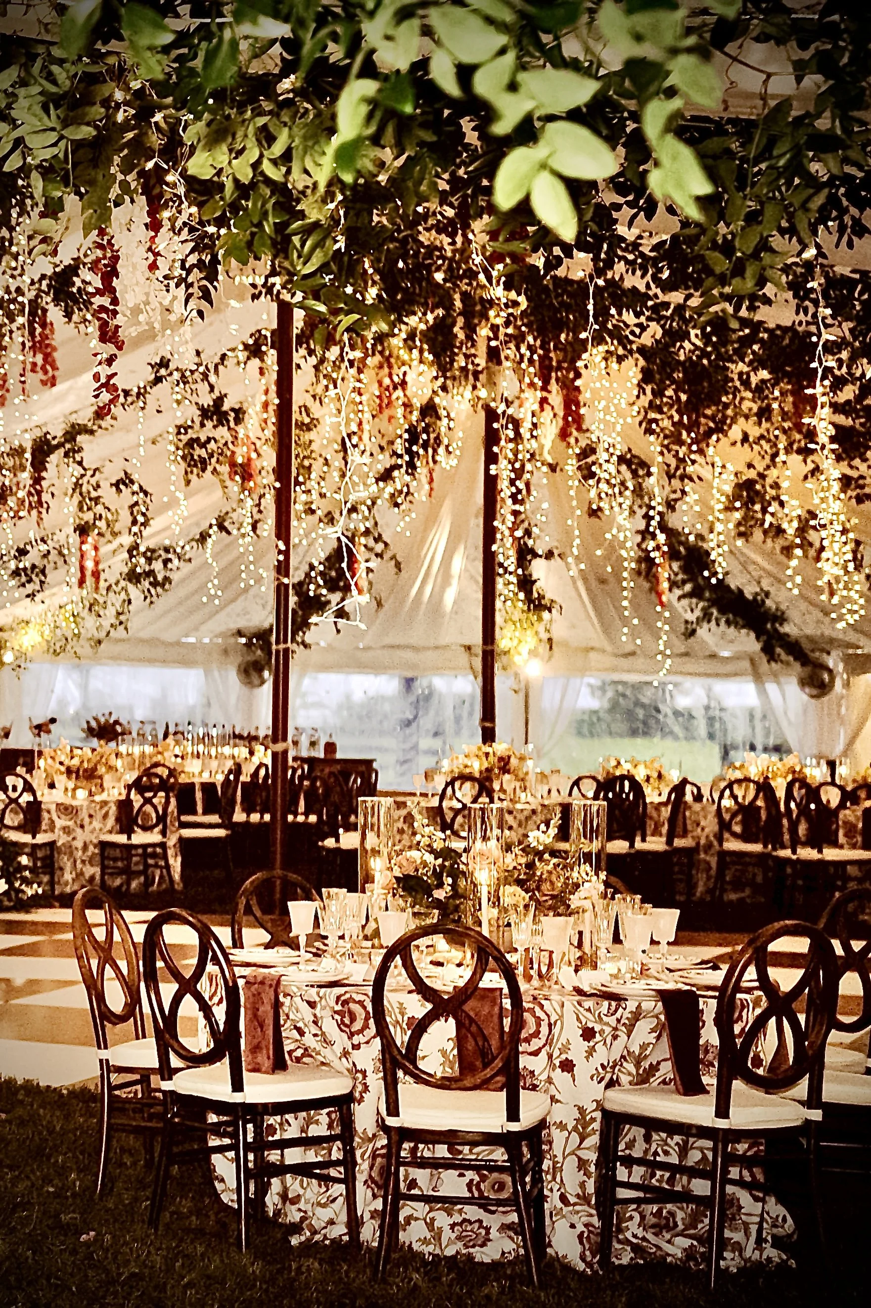 Elegant event setup inside a tent with decorated tables, floral centerpieces, and hanging string lights, creating a warm and festive atmosphere.