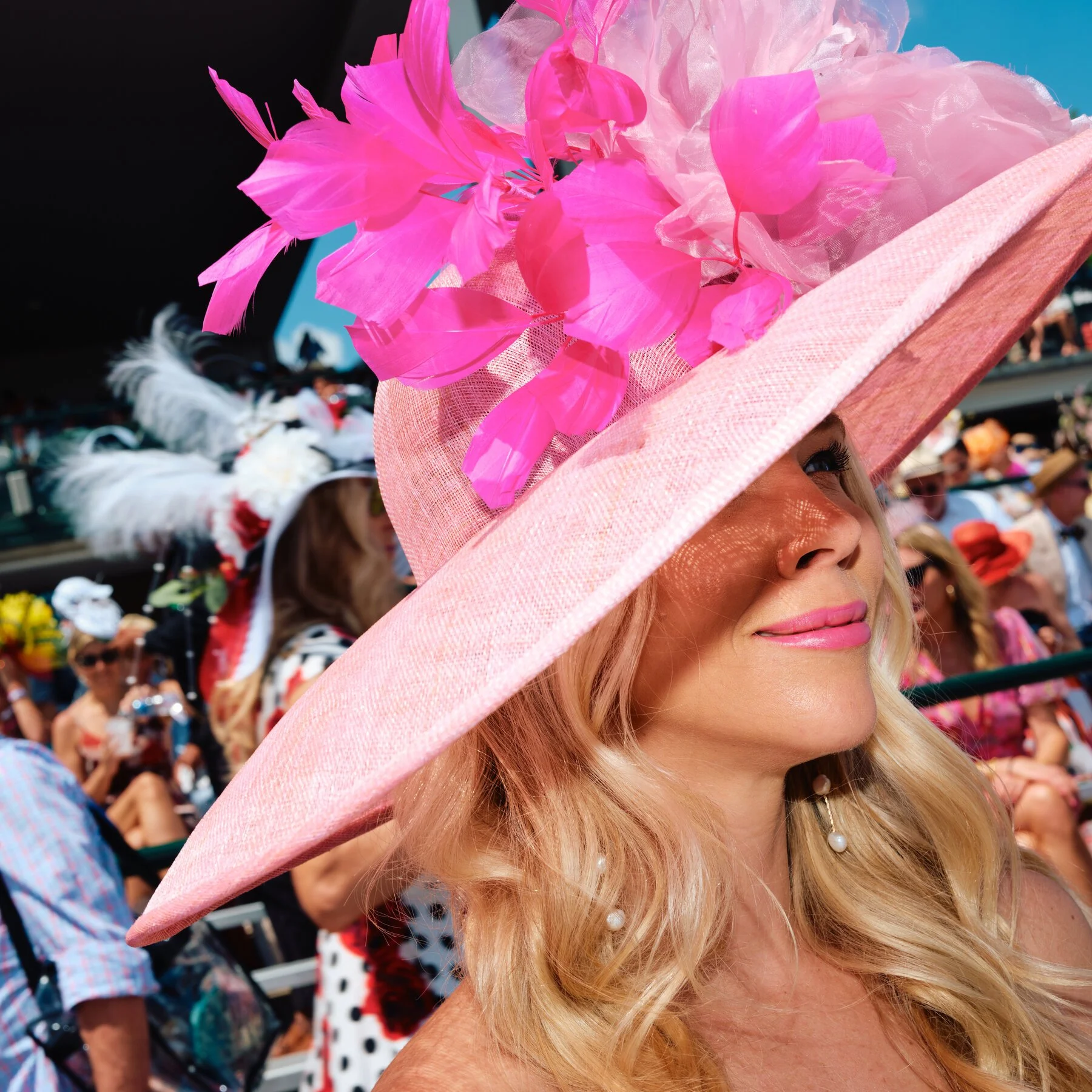 Woman wearing a large pink hat adorned with pink feathers for the Kentucky Oaks with a crowd in the background.