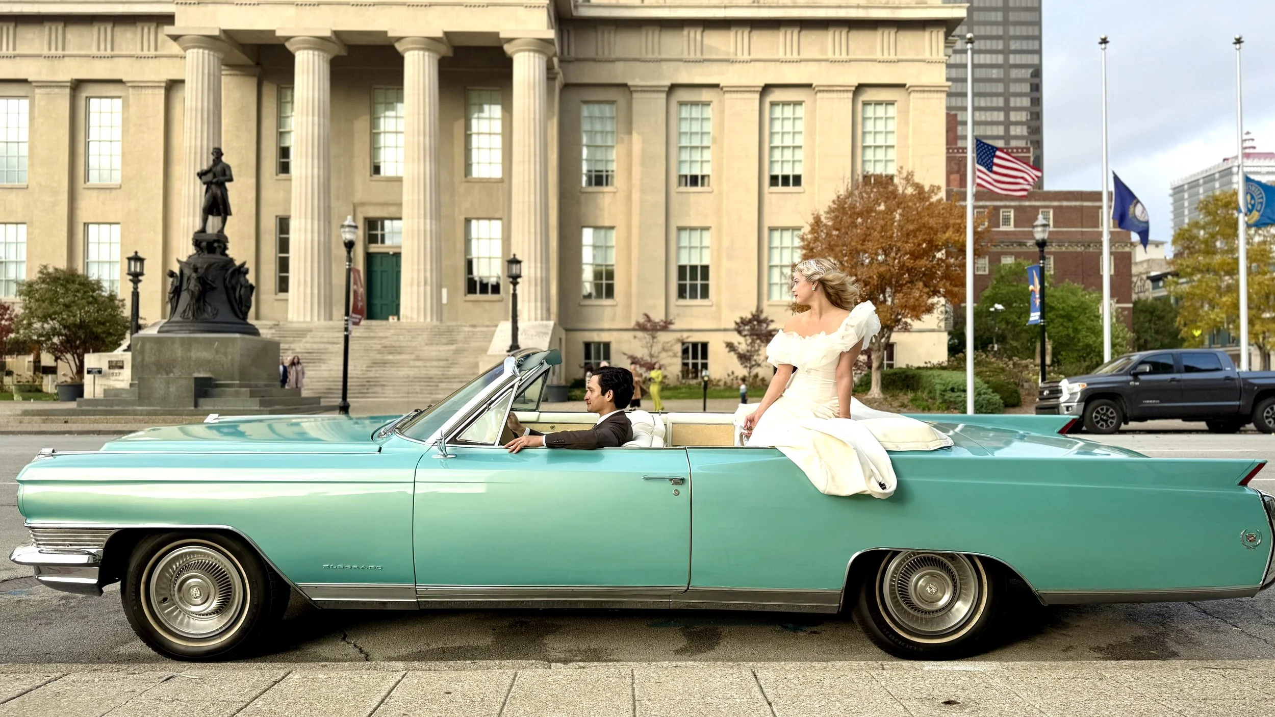 Close-up of the front end of Charlene, a 1964 Cadillac Eldorado convertible in Turino Turquoise with chrome details, parked outdoors with colorful buildings and a fountain in the background.