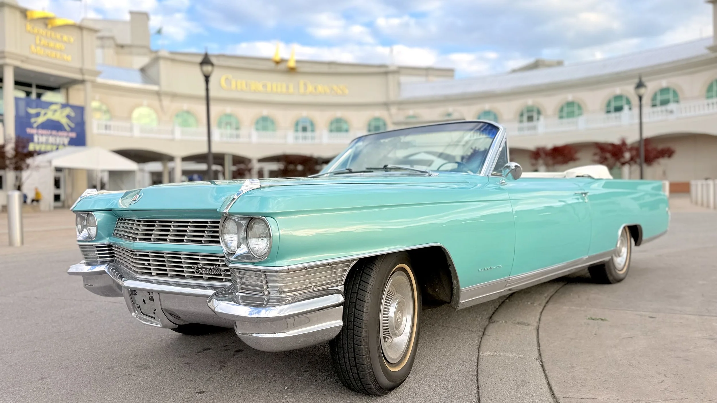 Charlene, a 1964 Cadillac Eldorado convertible in Turino Turquoise with chrome details, parked in the arrival circle with Churchill Downs in the background.