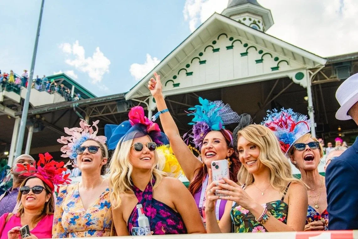Group of women at the Kentucky Derby, wearing colorful dresses and elaborate hats, taking a selfie and smiling.