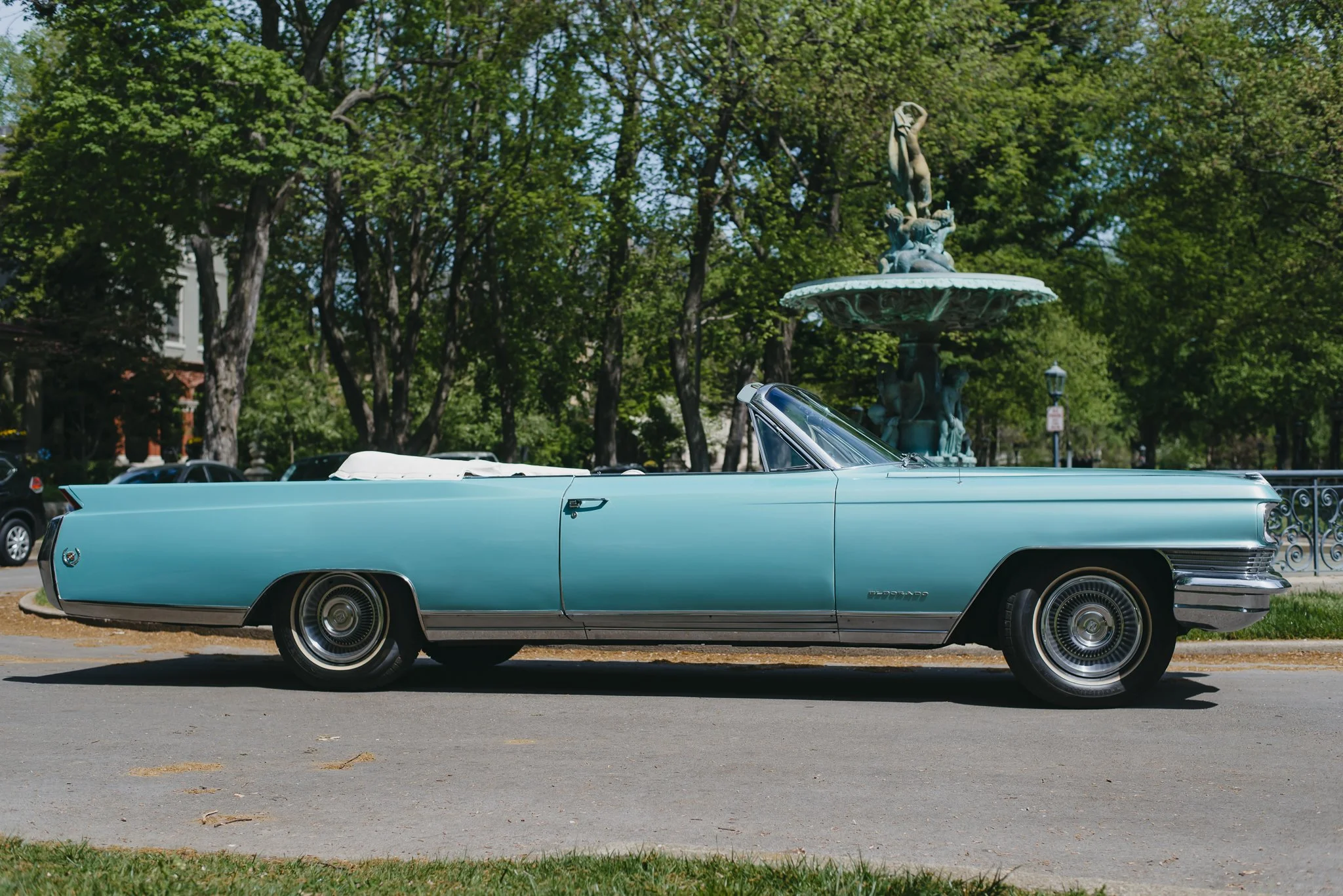 Charlene, a 1964 Cadillac Eldorado convertible, parked on a street with trees and a fountain in the background.
