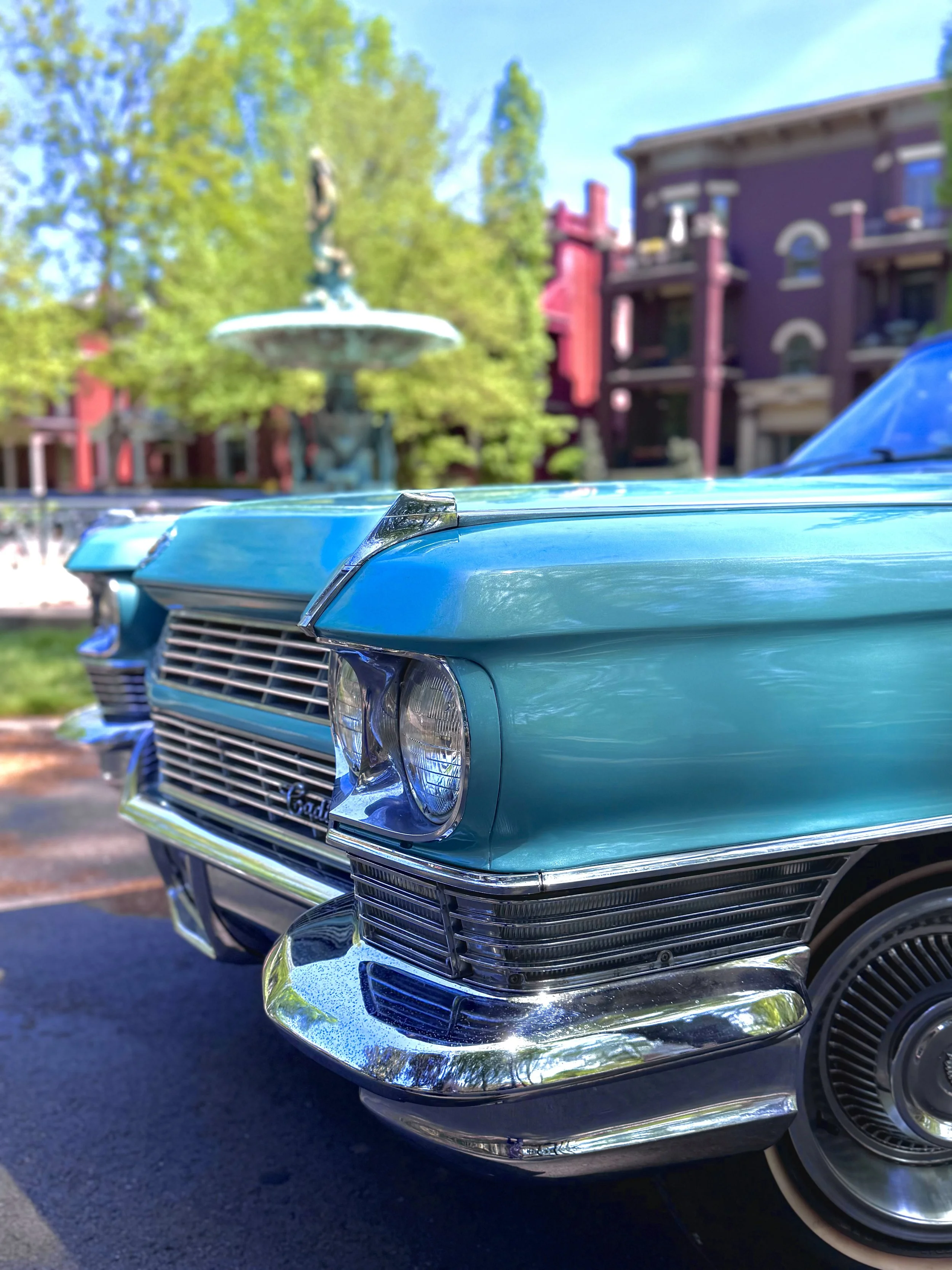 Close-up of the front end of Charlene, a 1964 Cadillac Eldorado convertible in Turino Turquoise with chrome details, parked outdoors with colorful buildings and a fountain in the background.