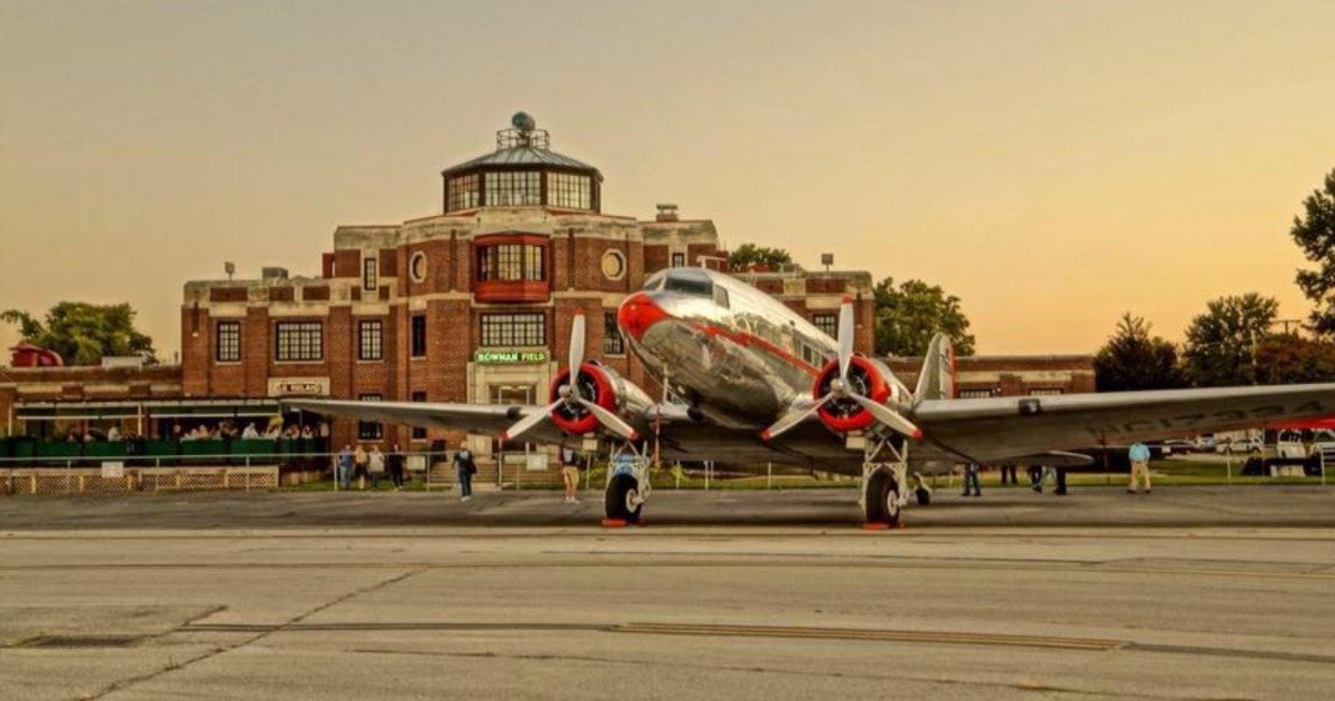 A vintage dual prop silver passenger airplane with red accents parked in front of an the historic terminal at Bowman Field in Louisville, KY at sunset.
