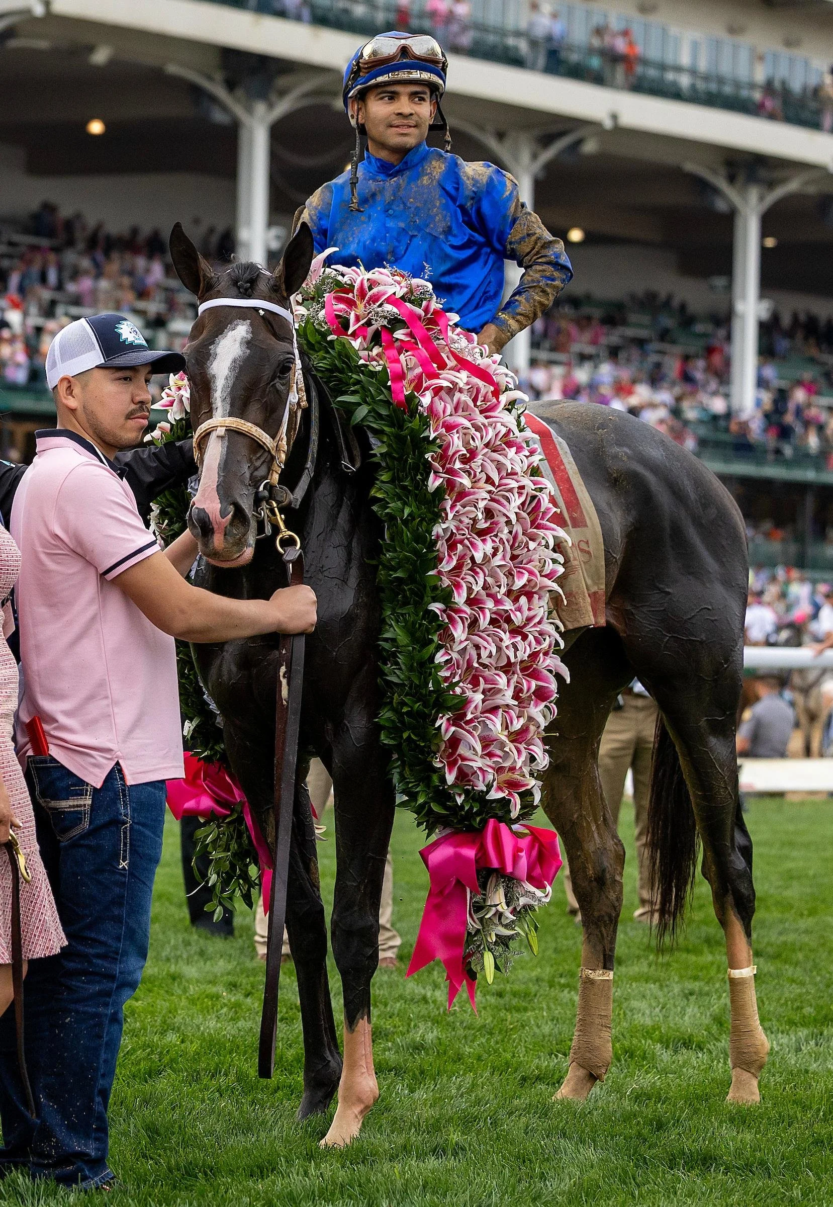 The winning Kentucky Oaks jockey sitting on the Oaks winner, decorated with a large lily garland at Churchill Downs, with spectators in the background.