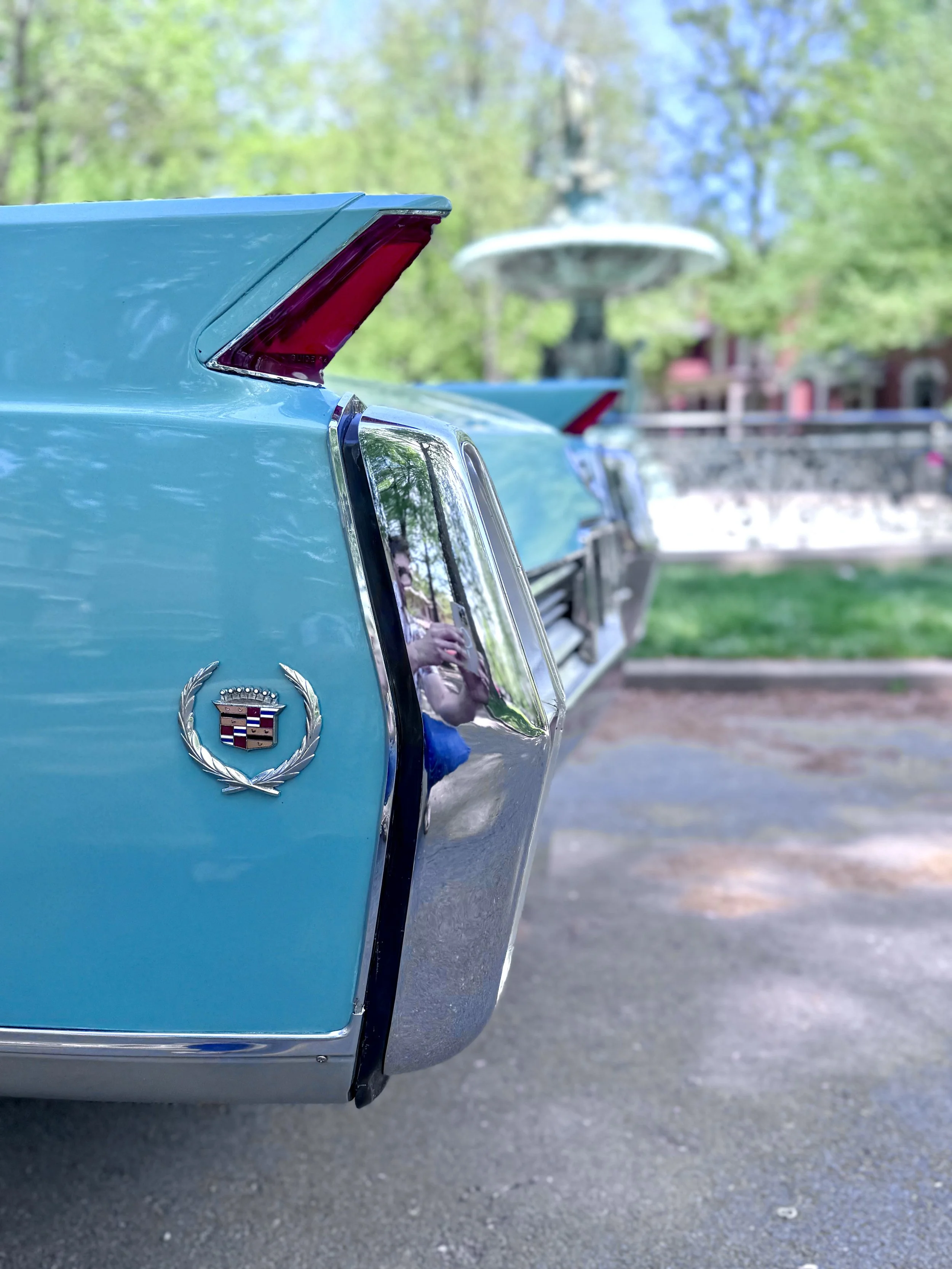 Close-up of the rear end of Charlene, a 1964 Cadillac Eldorado convertible in Turino Turquoise, showing the Cadillac emblem and tail light, with a park fountain and trees in the background.
