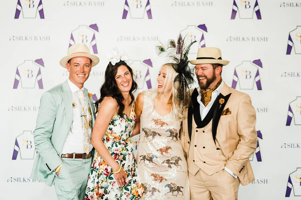 Four people on the purple carpet dressed in Derby best, smiling and standing close together in front of a backdrop with logos and text reading 'Silks Bash.'