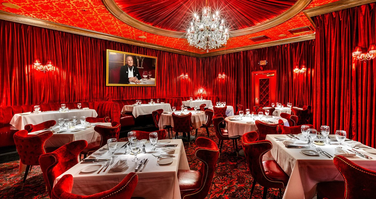 The Ruby Room at Jeff Ruby's, Louisville, with red velvet curtains, red chairs, and white tablecloths set for dining. A chandelier hangs from the ceiling, and there is a large portrait of a man in formal attire on the wall.
