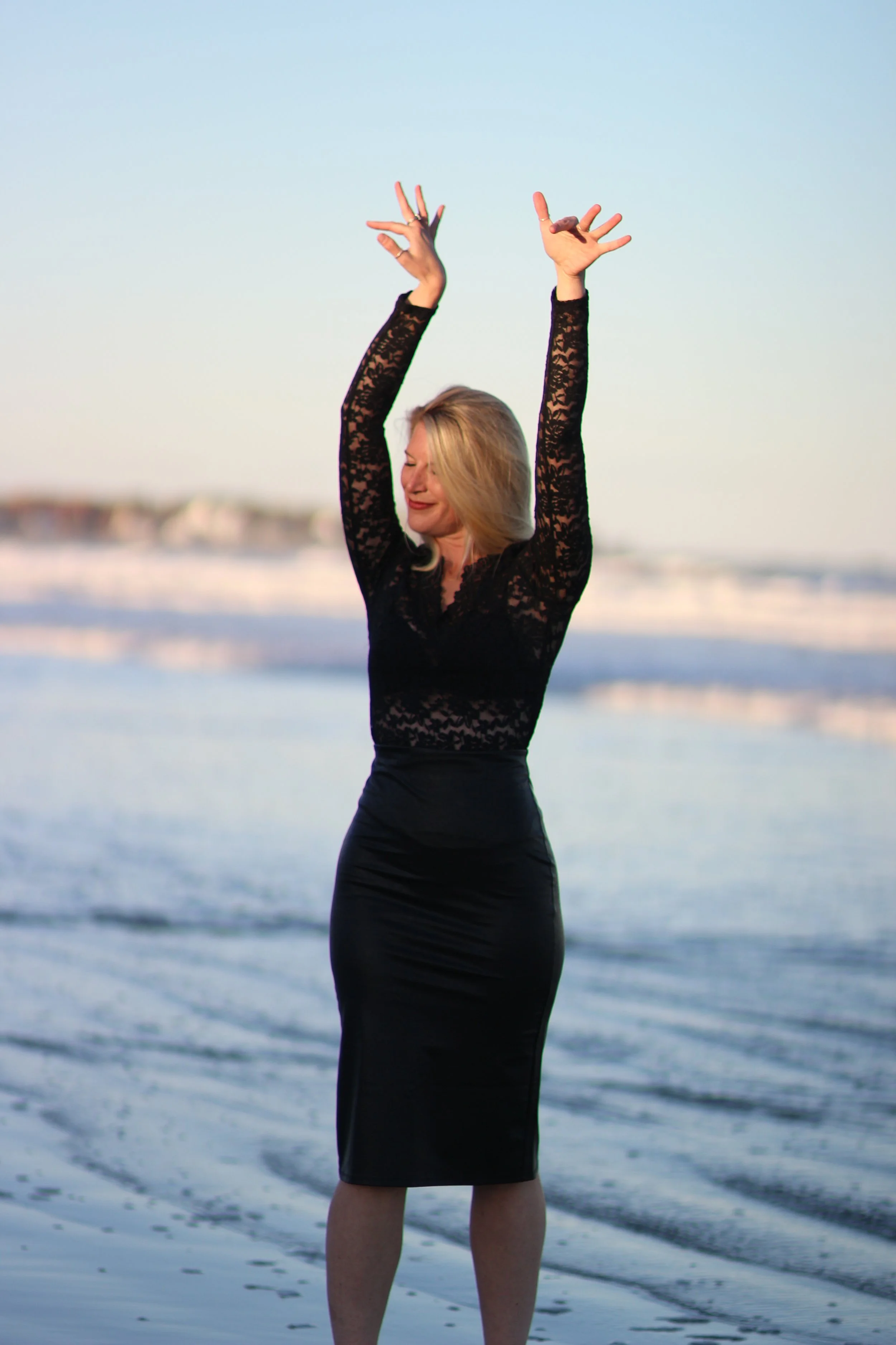 A woman with blonde hair wearing a black lace top and black skirt dancing or stretching on the beach with her arms raised, near the water during sunset or sunrise.