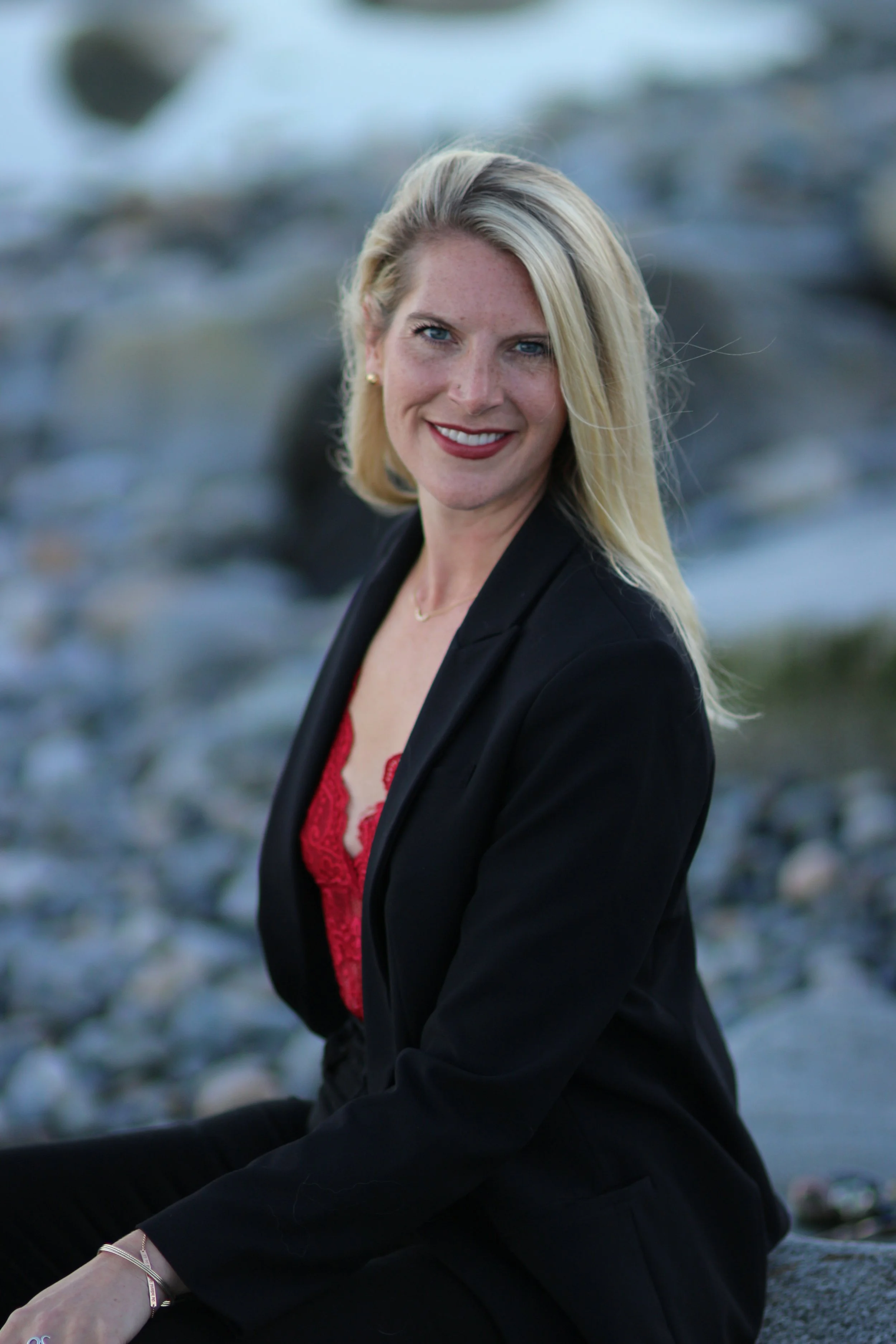 A woman with blonde hair smiling while sitting on rocks by a river, wearing a black blazer and red lace top.