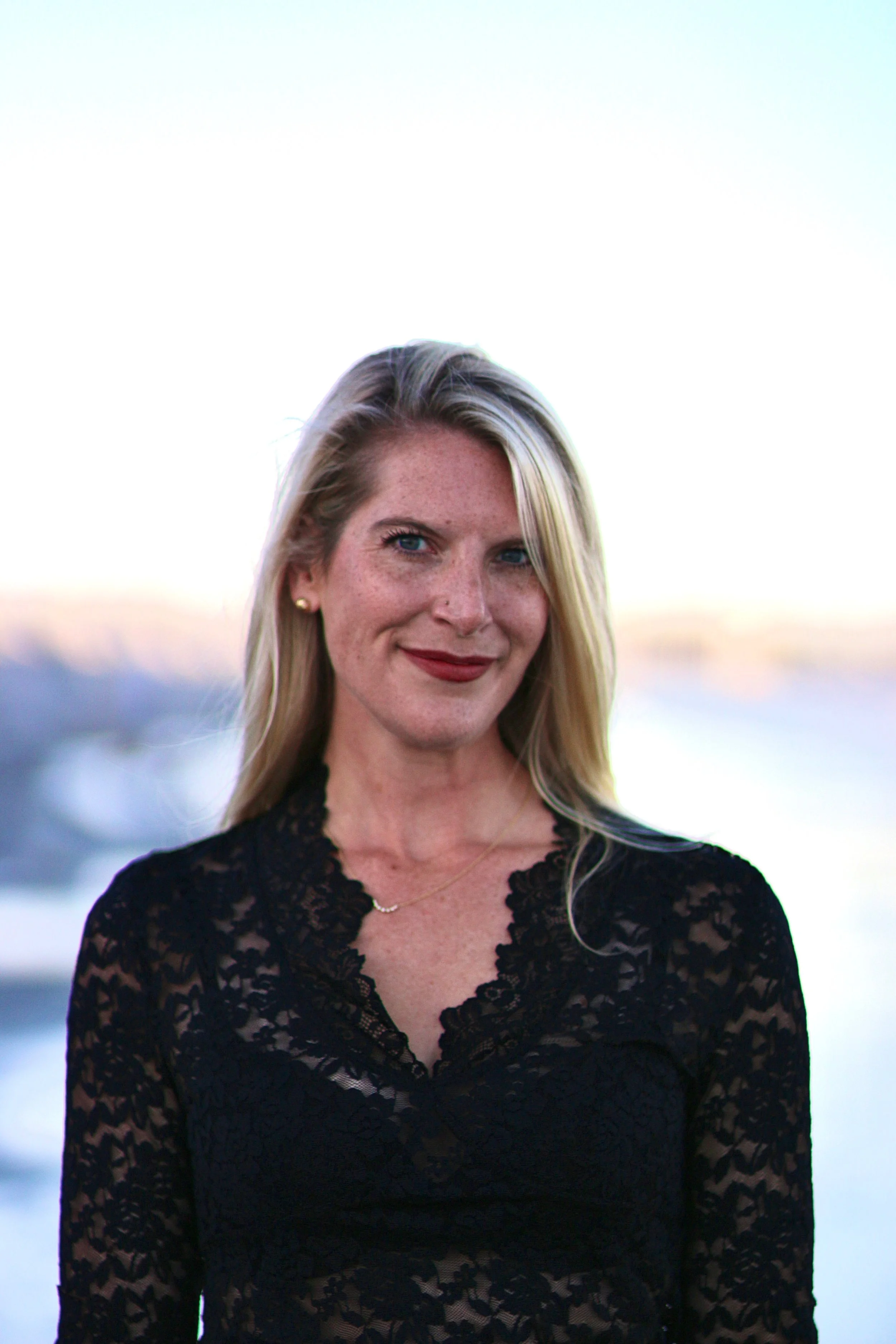 A woman with blonde hair, blue eyes, wearing a black lace top, standing outdoors near water during sunset.