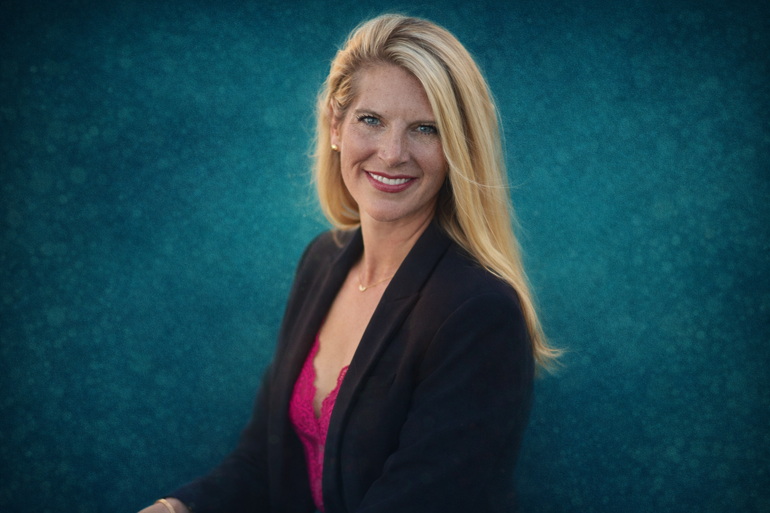 Portrait of a smiling blonde woman in a black blazer with a pink lace top, standing against a textured teal background.