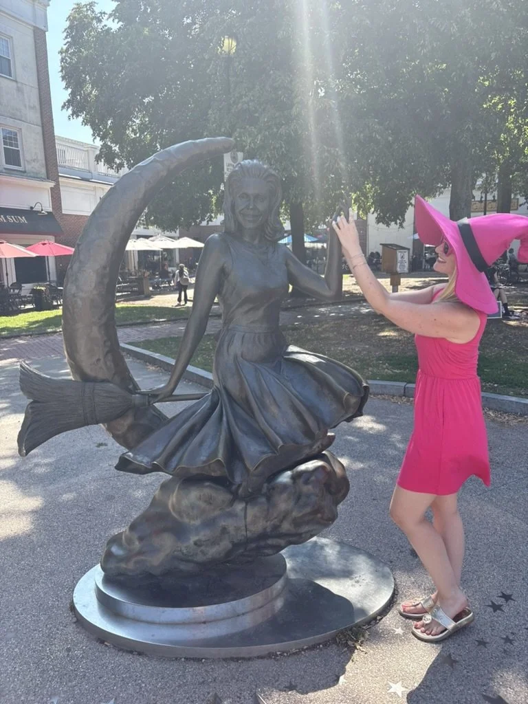 A woman in a pink dress and large pink sunhat appears to be giving a high five to a statue of a girl in a dress sitting on a fish, part of a fountain sculpture.