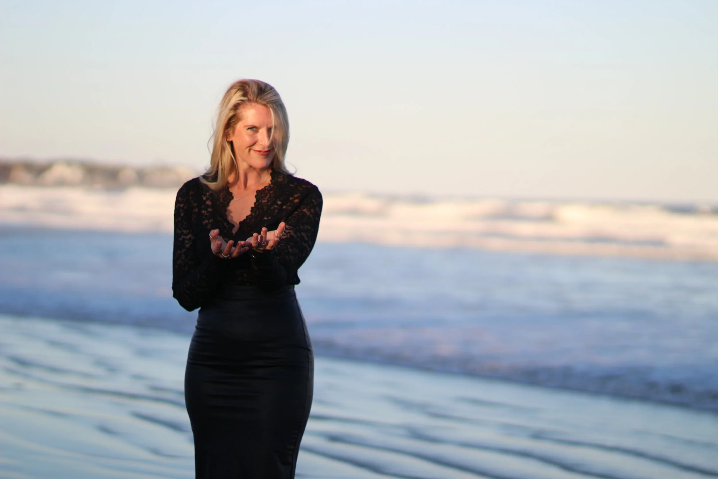 A woman with blonde hair wearing a black lace top and a black skirt standing on a beach with waves in the background during sunset or sunrise, looking towards the camera and smiling.