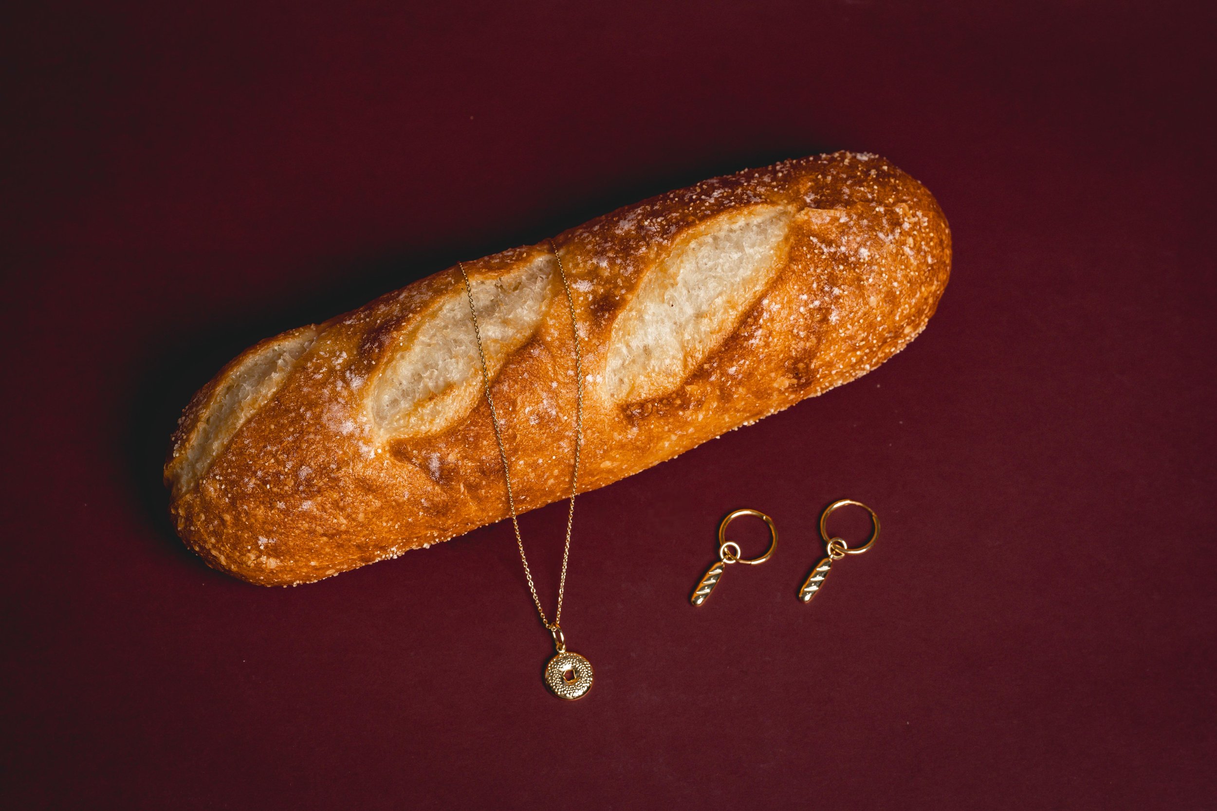 A loaf of bread with a jewelry necklace and a pair of small hoop earrings with feather charms on a dark red background.