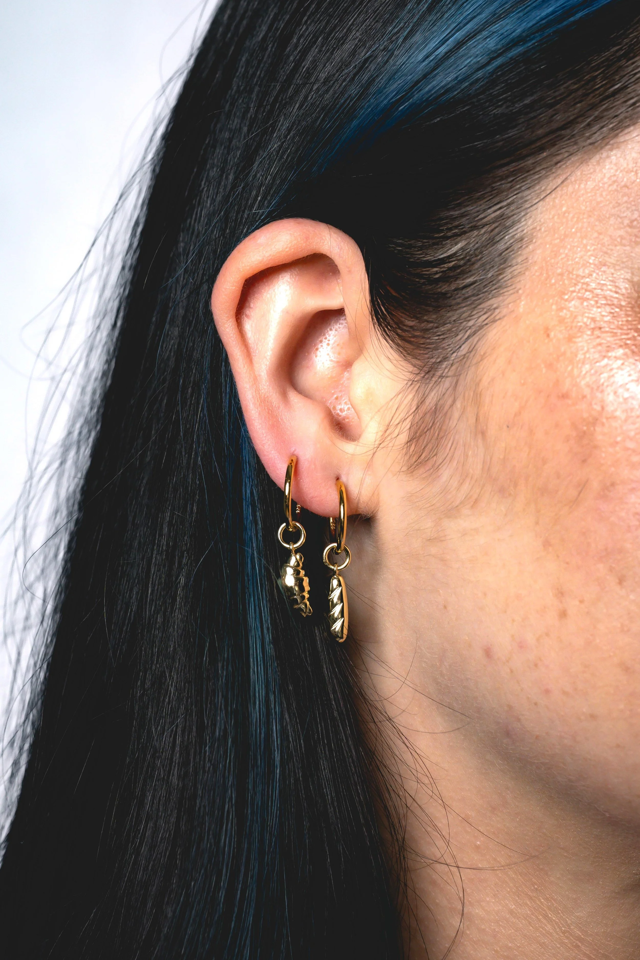 Close-up of a woman's ear with two gold hoop earrings, one with a small charm. The woman has black hair with blue highlights.
