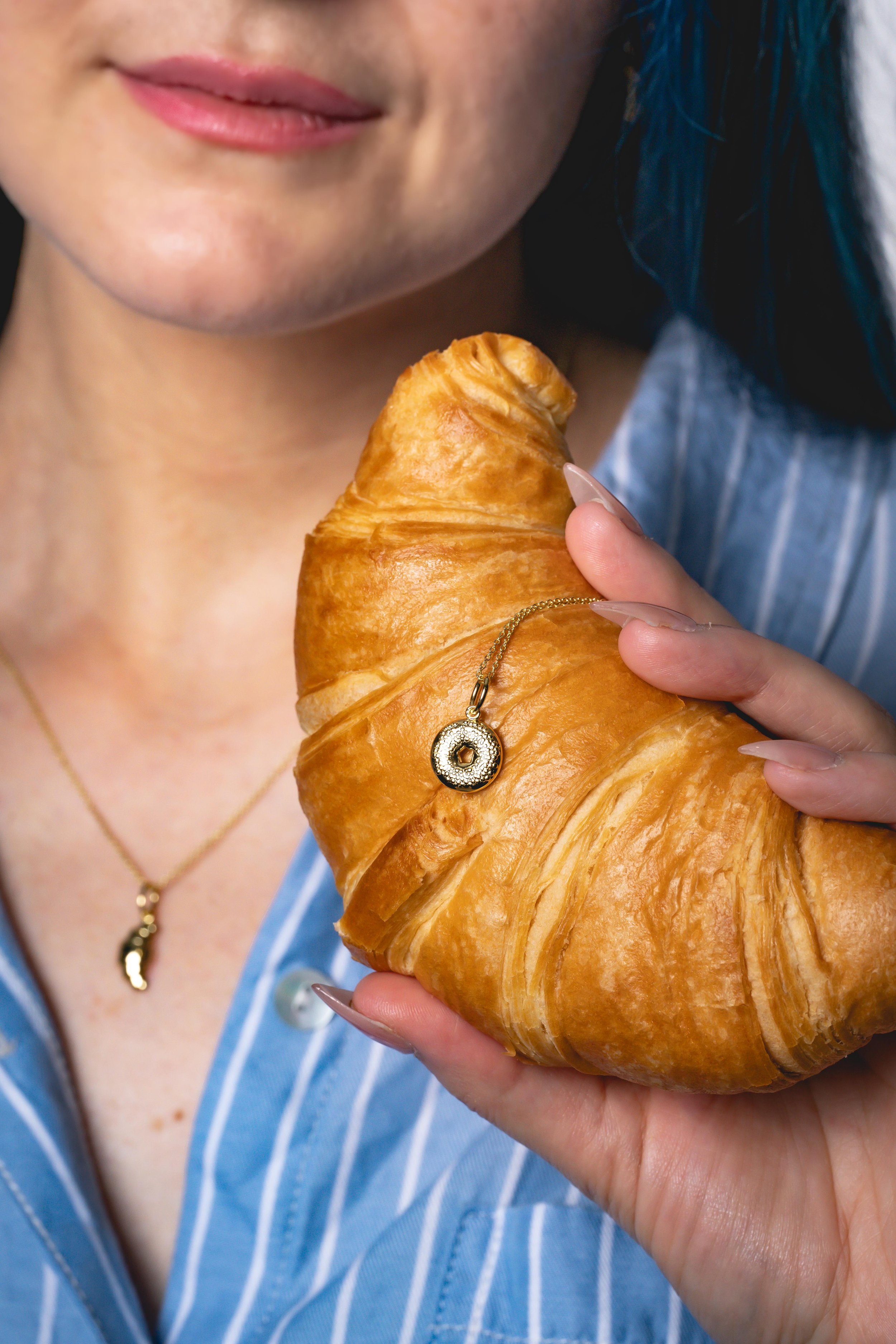 A woman holding a large croissant with a gold necklace featuring a donut pendant, wearing a blue striped shirt.