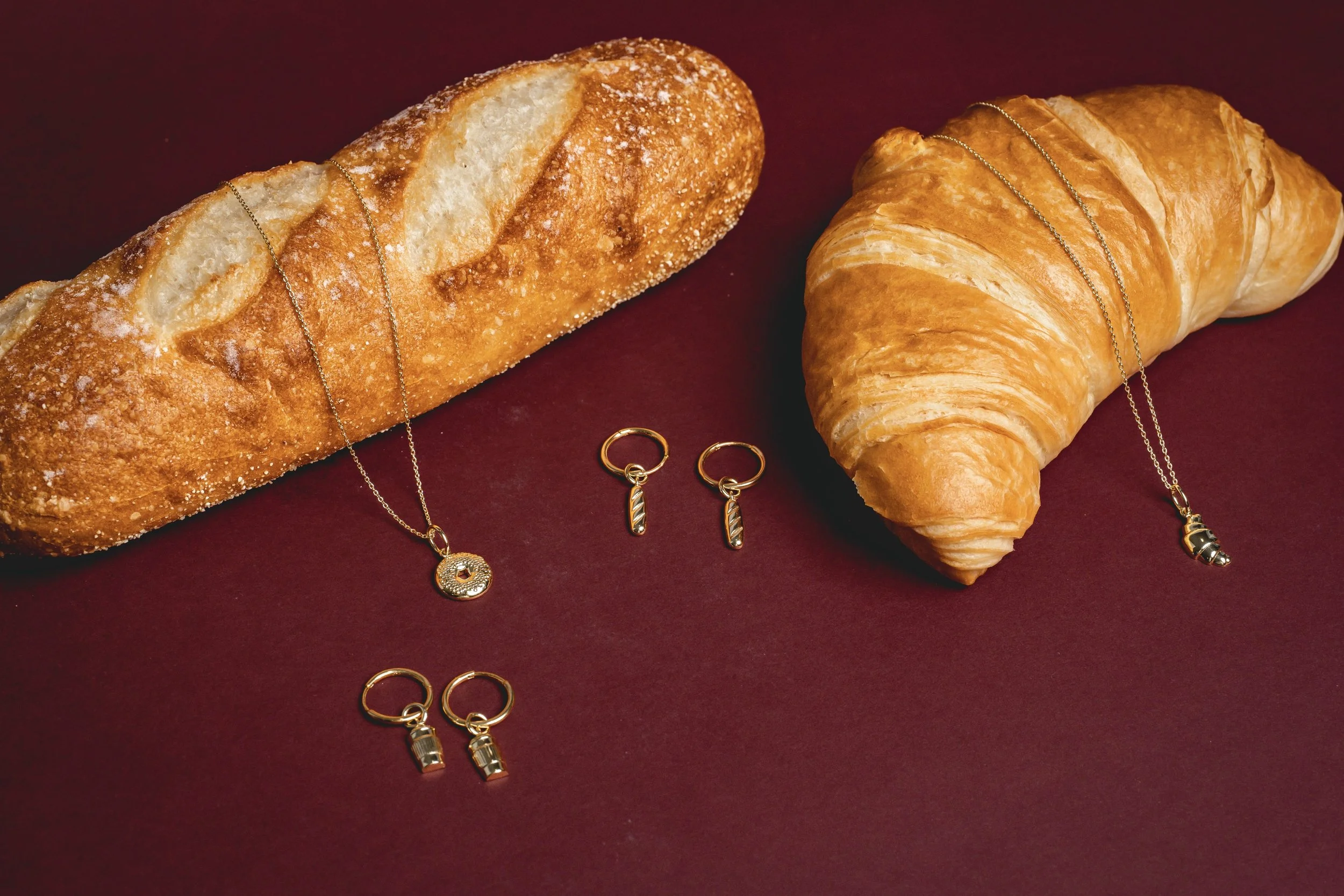 Golden necklaces and earrings arranged on a burgundy surface, with a baguette and a croissant in the background.