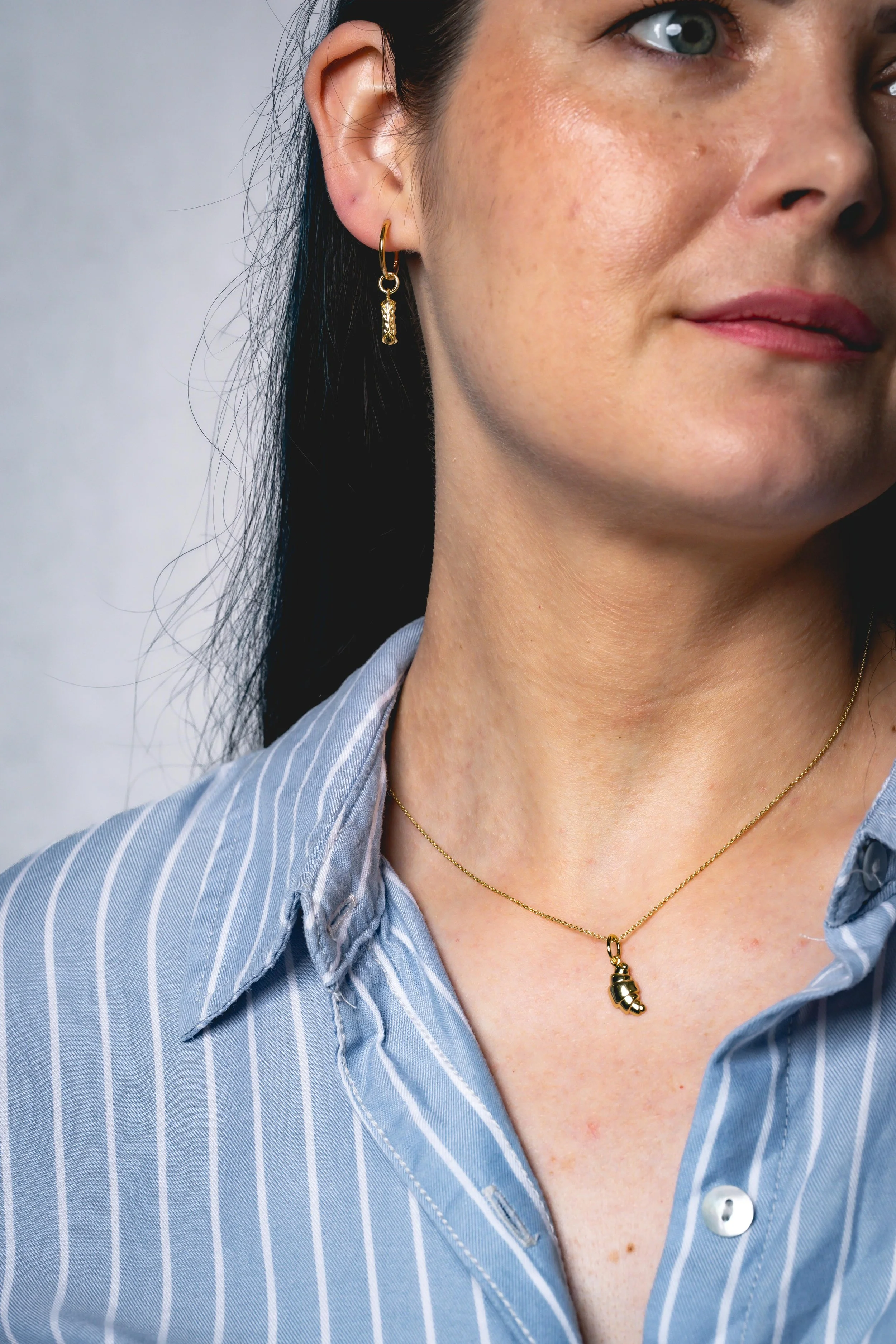 Close-up of a woman's face and neck, showing her wearing gold jewelry, including a small hoop earring and a necklace with a pendant, along with a light blue and white striped collared shirt.