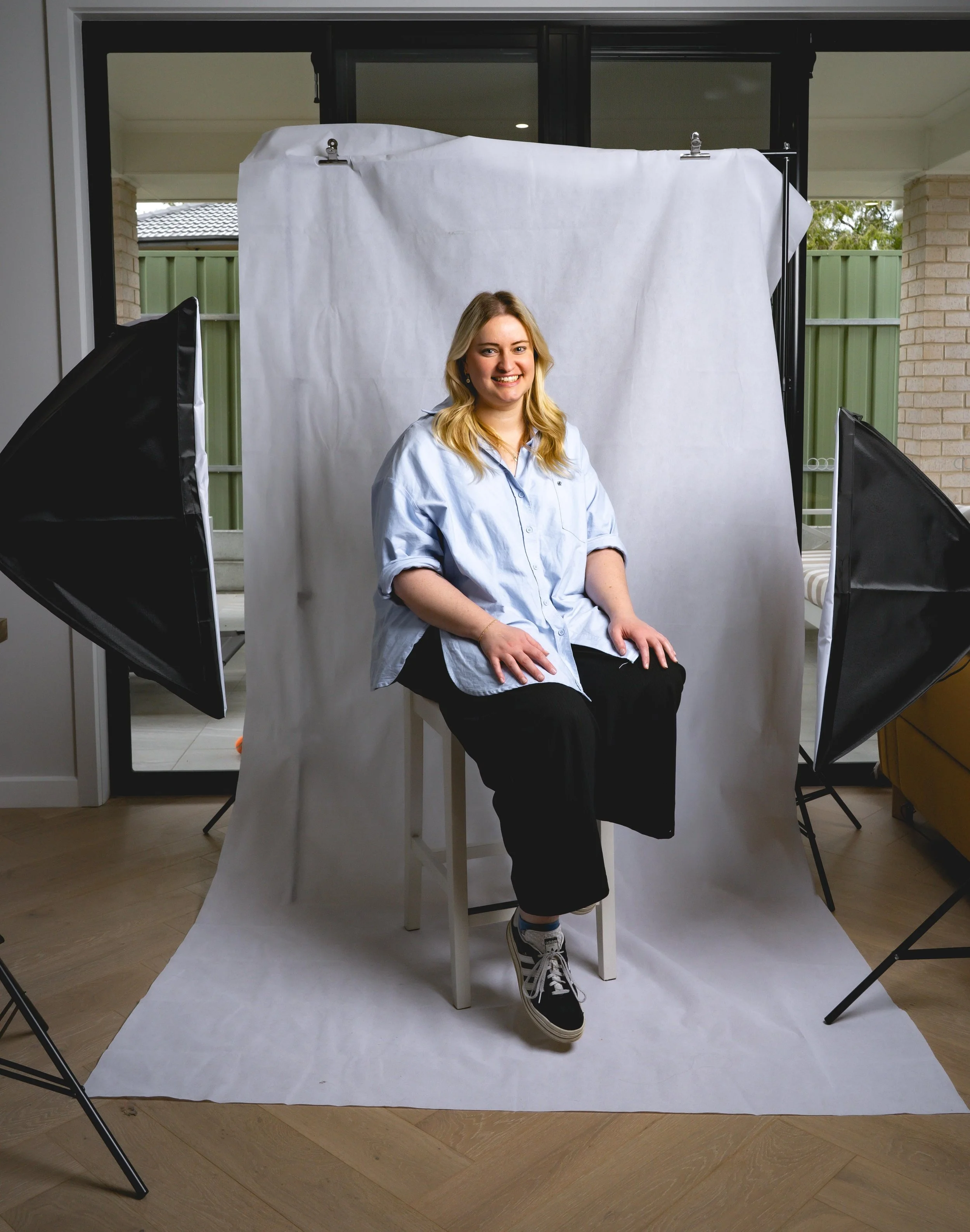 A young woman sitting on a stool during a photo shoot in a home studio with softbox lights and a white backdrop.