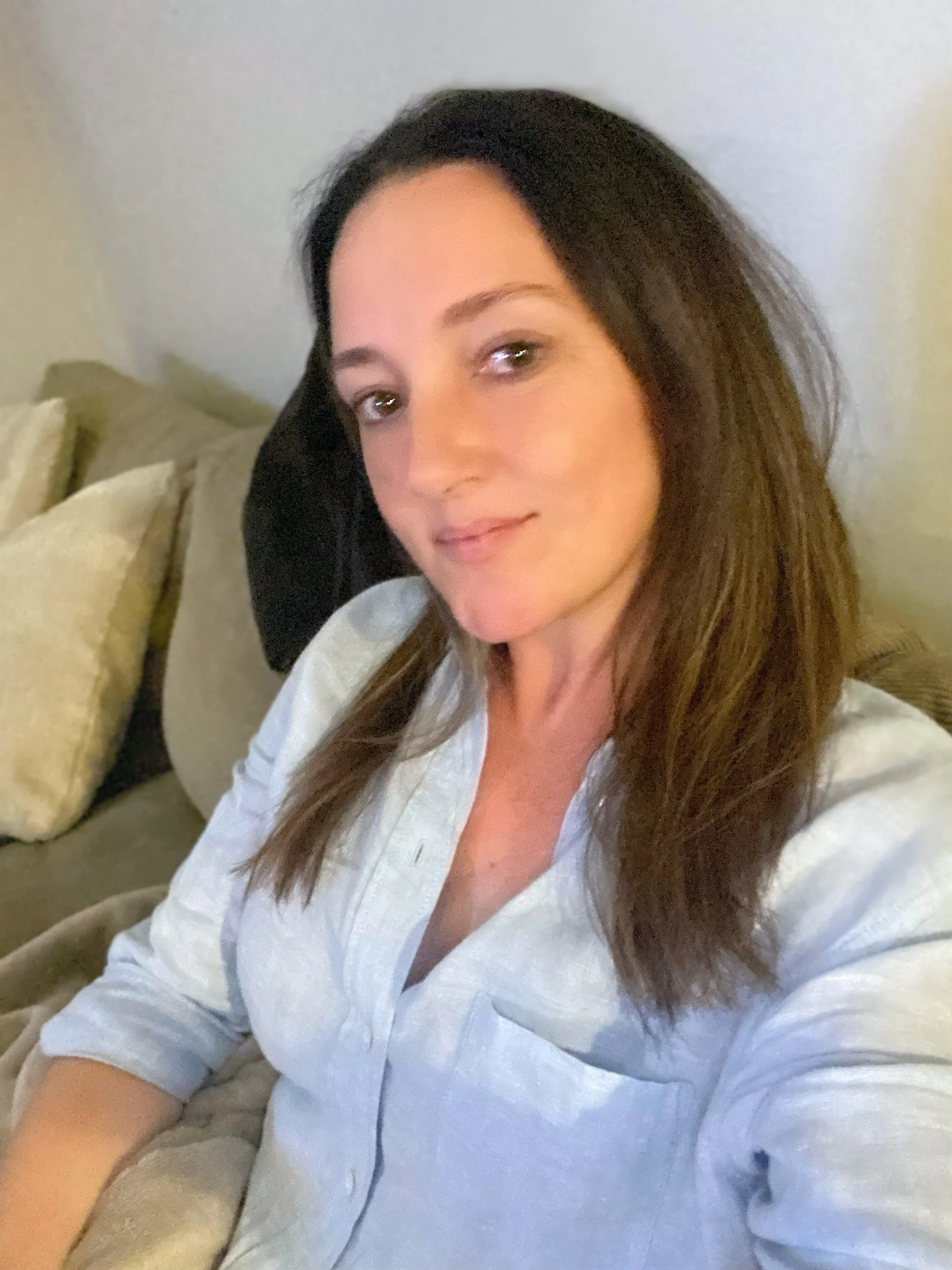 A woman with shoulder-length brown hair and light skin, wearing a white shirt, sitting on a beige lounge with multiple pillows in a cozy indoor setting. Copyright M Thornton