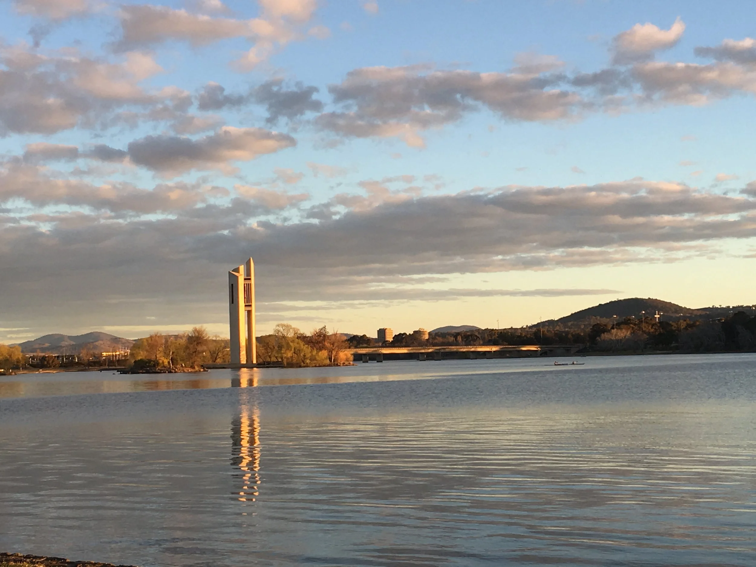 Lake Burley Griffin in Canberra, with calm water reflecting the sky and a tall, modern bridge structure with a tower (The Carillion), with hills and trees in the background under a partly cloudy sky at sunset. Copyright M Thornton