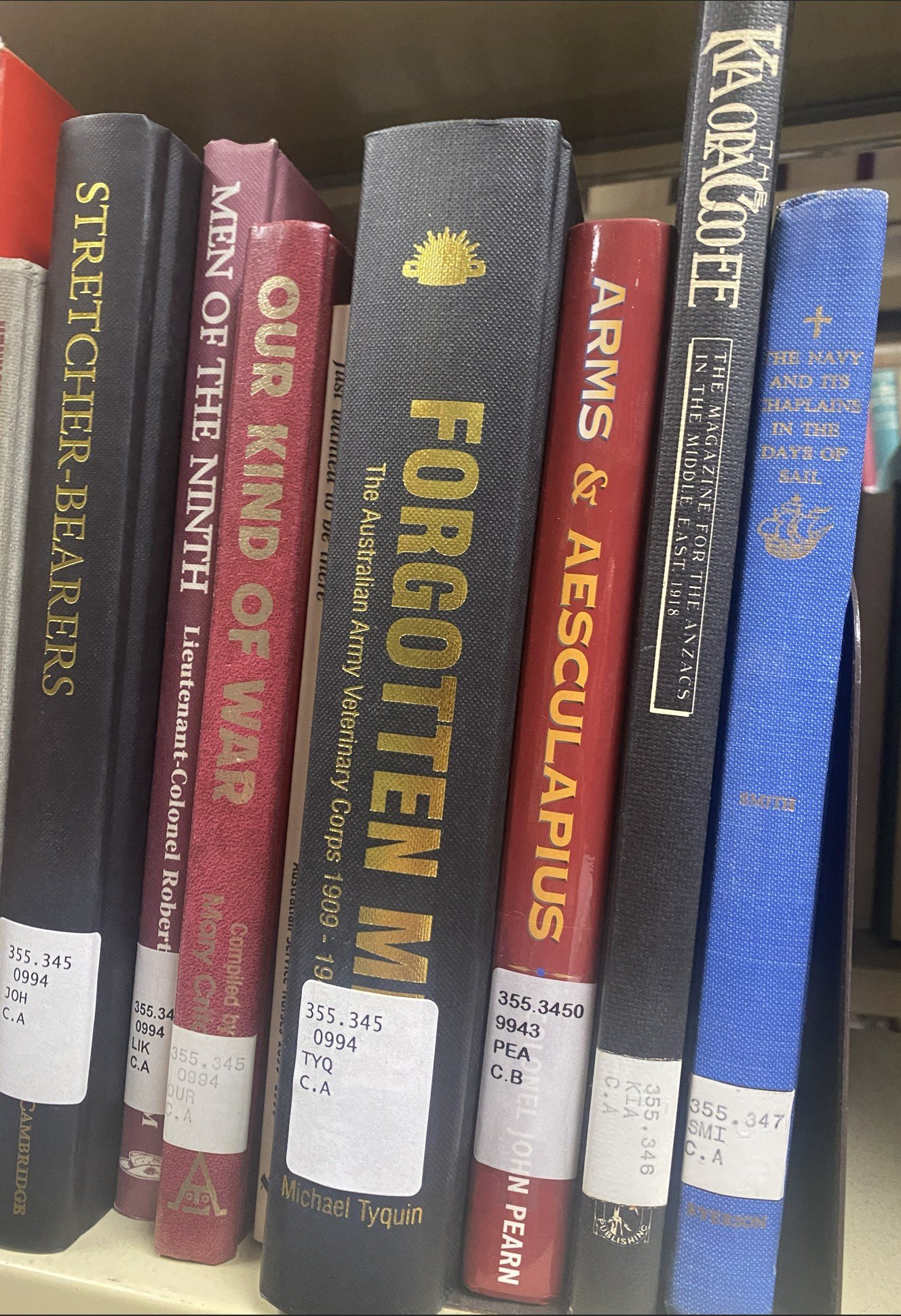 A row of books on a library shelf, including titles about Australian army Vietnam groups, a children's book, and military history.