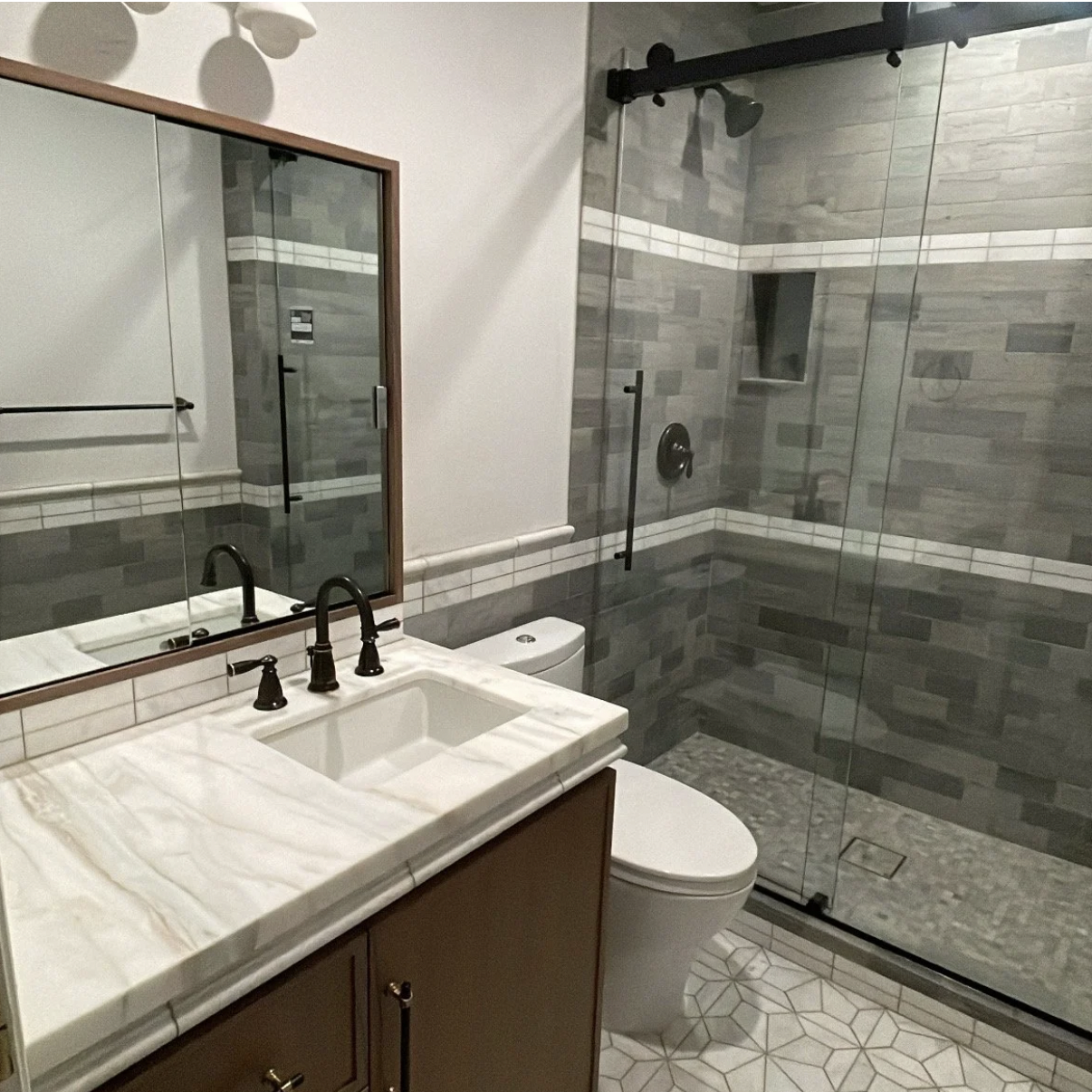 Modern bathroom with a glass-enclosed shower, white vanity with black fixtures, a mirror, and a toilet. The shower has gray subway tiles and a recessed shelf.