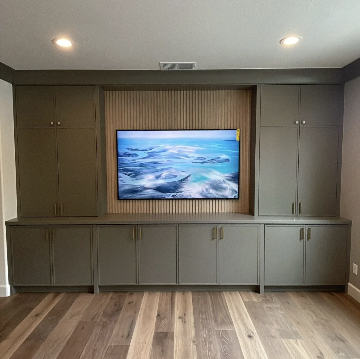 Built-in entertainment center with a flat-screen TV showing an aerial view of icy landscape, grey cabinetry on either side, and wood paneling behind the TV.