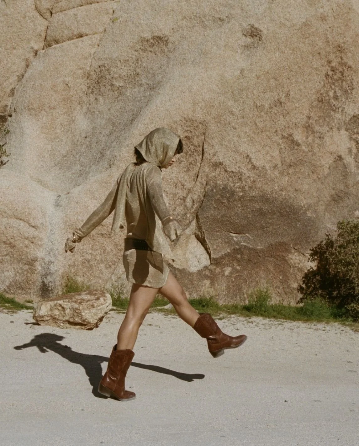 Woman walking on sandy terrain with a large rock formation and some greenery in the background, wearing a hoodie, shorts, and brown boots.