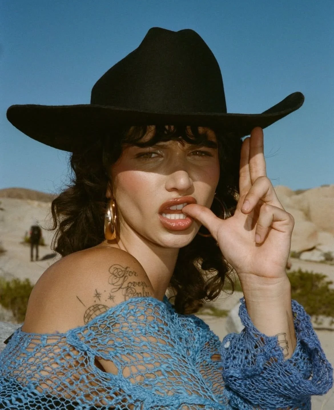 A woman wearing a large black hat, gold hoop earrings, a blue crocheted top, and makeup, with tattoos on her shoulder and arm, making a playful pose with her finger near her mouth in front of a desert landscape.