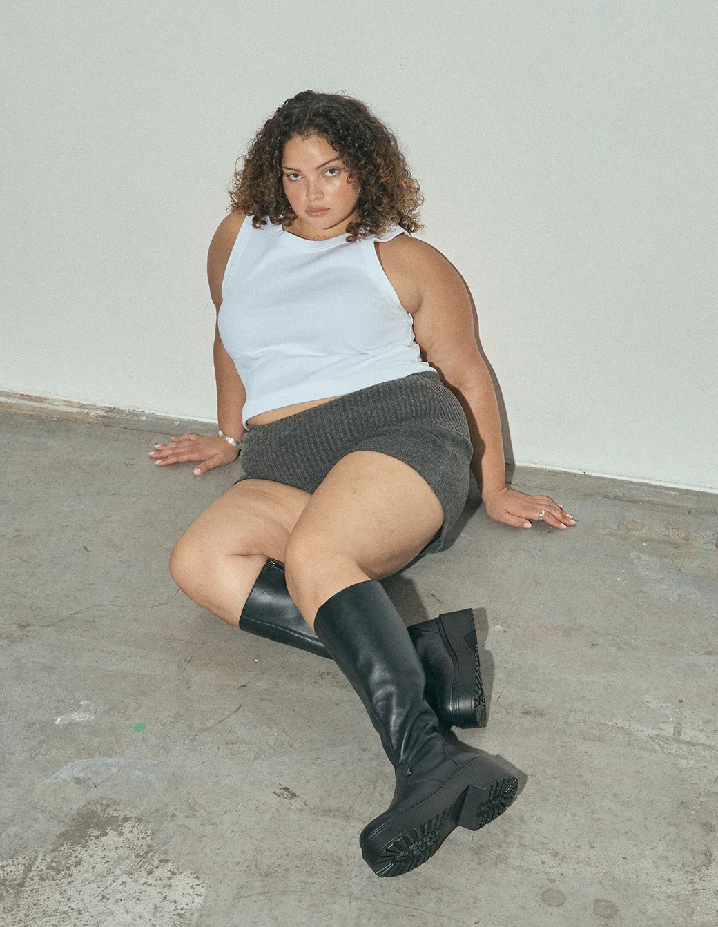 A woman with curly hair is sitting on a concrete floor against a plain white wall, wearing a white sleeveless top, dark shorts, and tall black boots, looking directly at the camera.