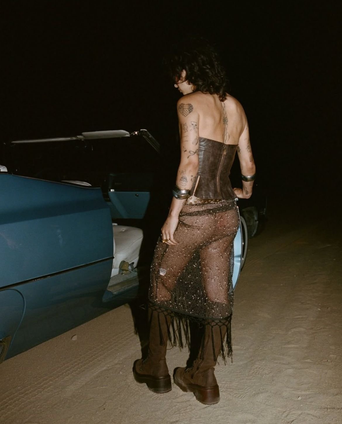 A woman with tattoos is standing next to a vintage car at night, wearing a corset, fishnet skirt, and boots.
