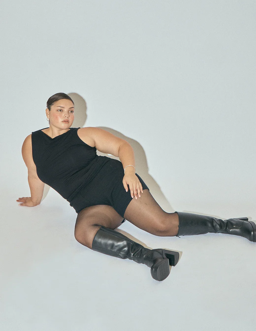 A woman in a black sleeveless top and black skirt sitting on the floor with high-heeled knee-high boots, against a plain white background.