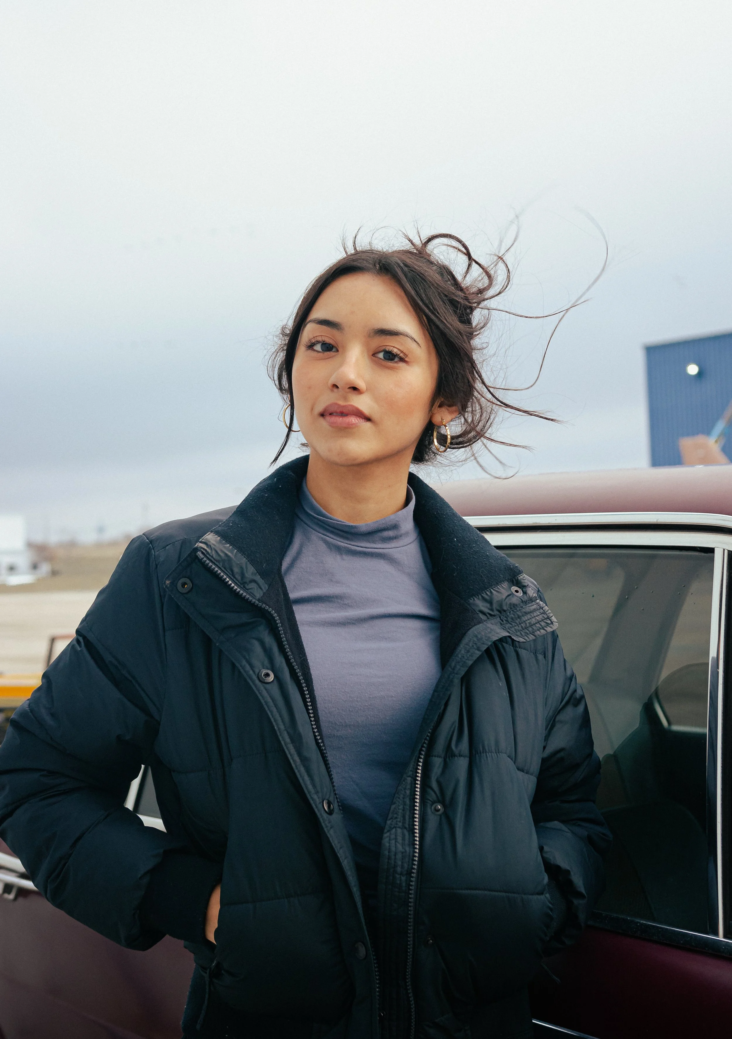 A young woman with short brown hair and hoop earrings stands outside next to a vintage car, wearing a black puffy jacket and a gray turtleneck, with a cloudy sky in the background.