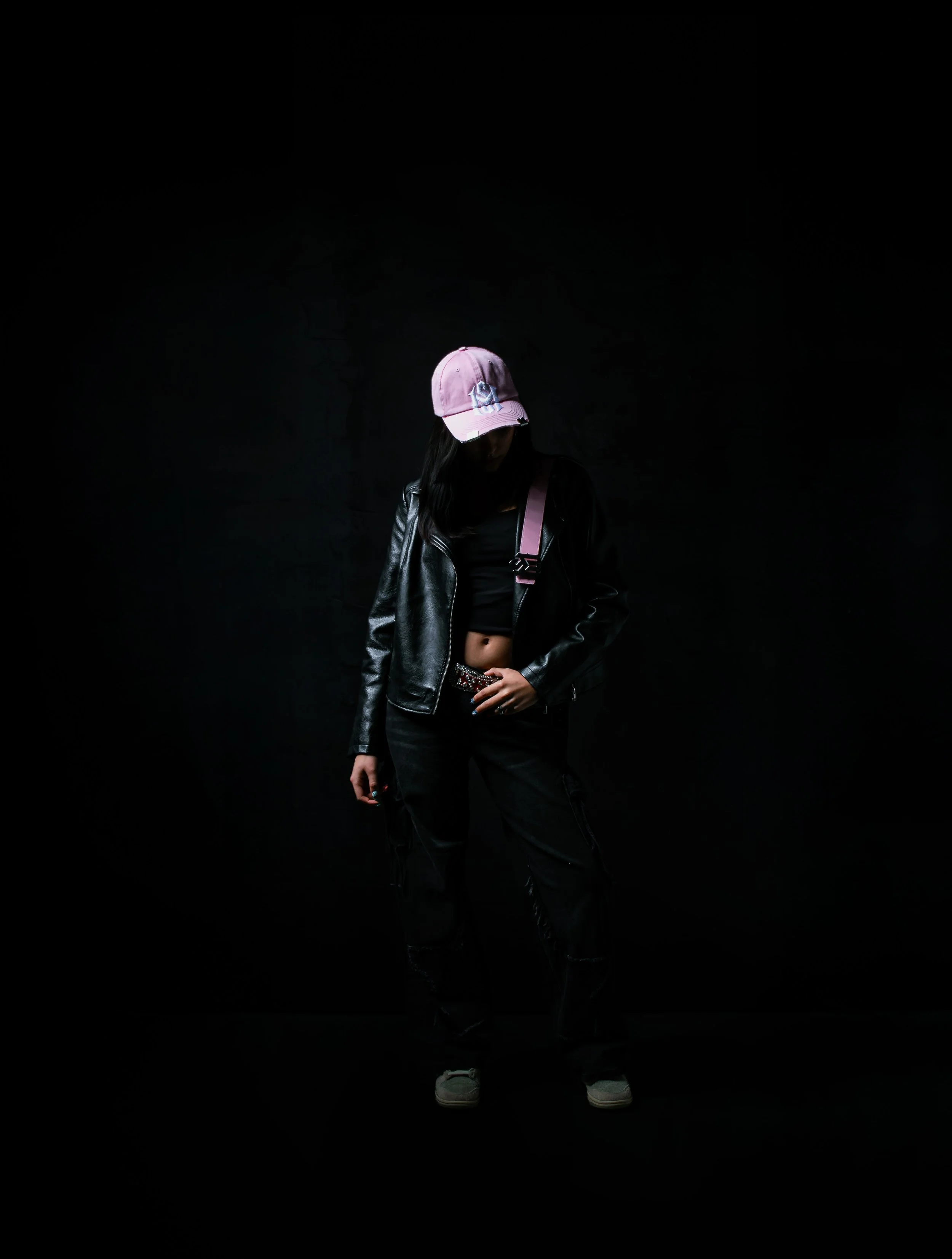 A person standing against a black background, wearing a pink cap, black leather jacket, black pants, and grey shoes, with a pink strap over their shoulder.